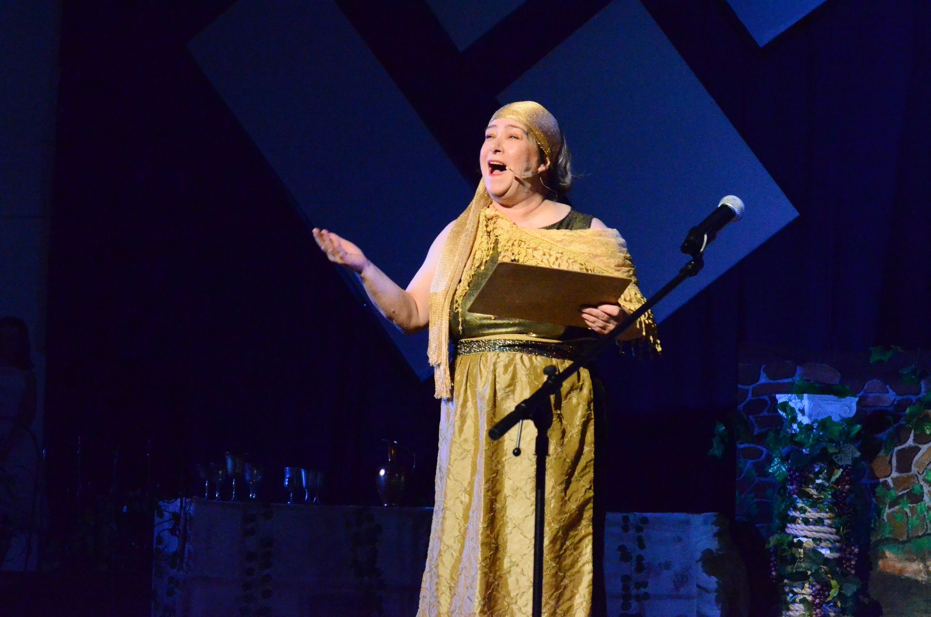 A performer in a gold costume and shawl stands at a microphone on stage, holding a script and gesturing expressively against a backdrop of geometric blue panels and scenic elements.