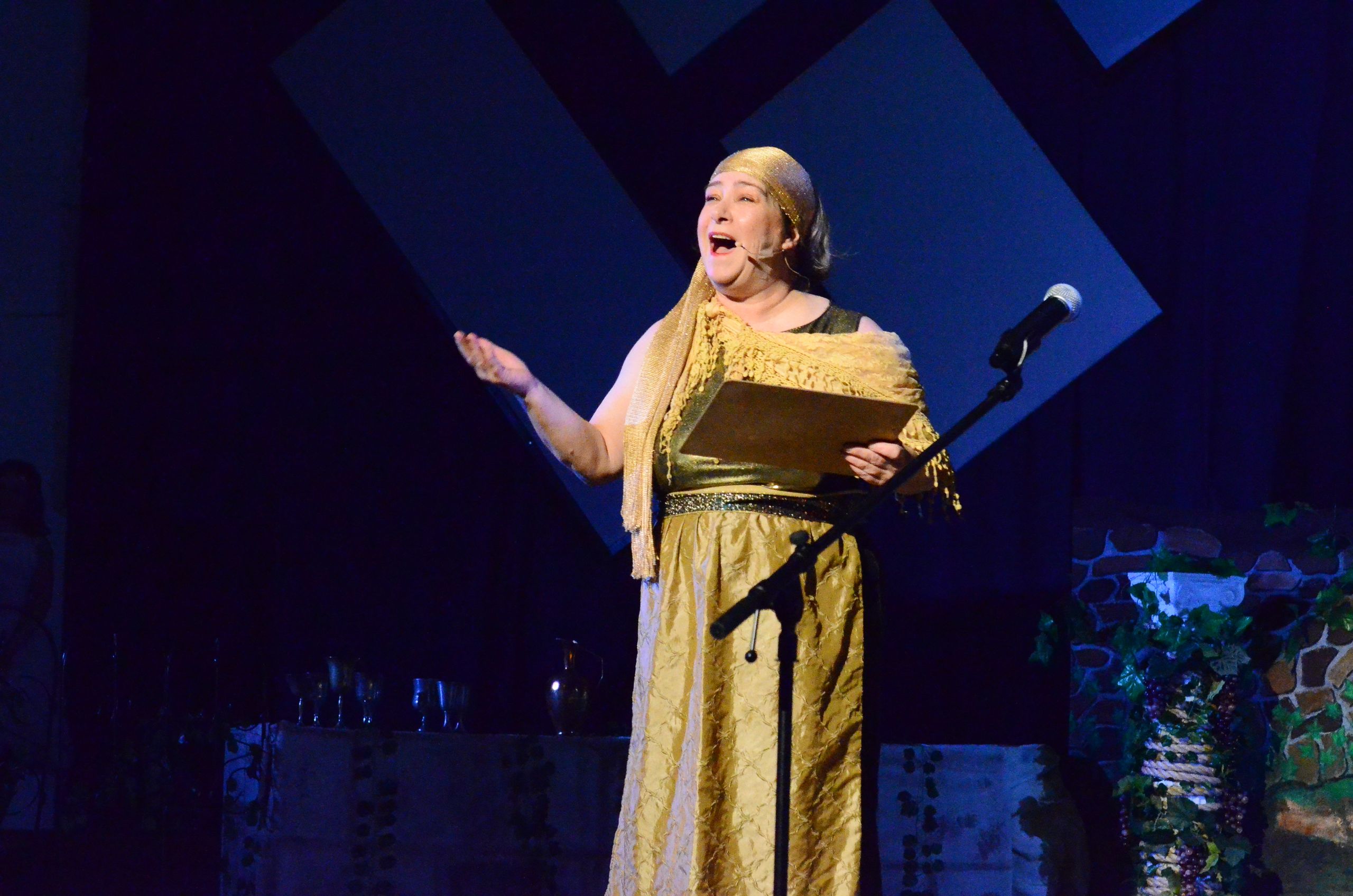 A performer in a gold costume and shawl stands at a microphone on stage, holding a script and gesturing expressively against a backdrop of geometric blue panels and scenic elements.