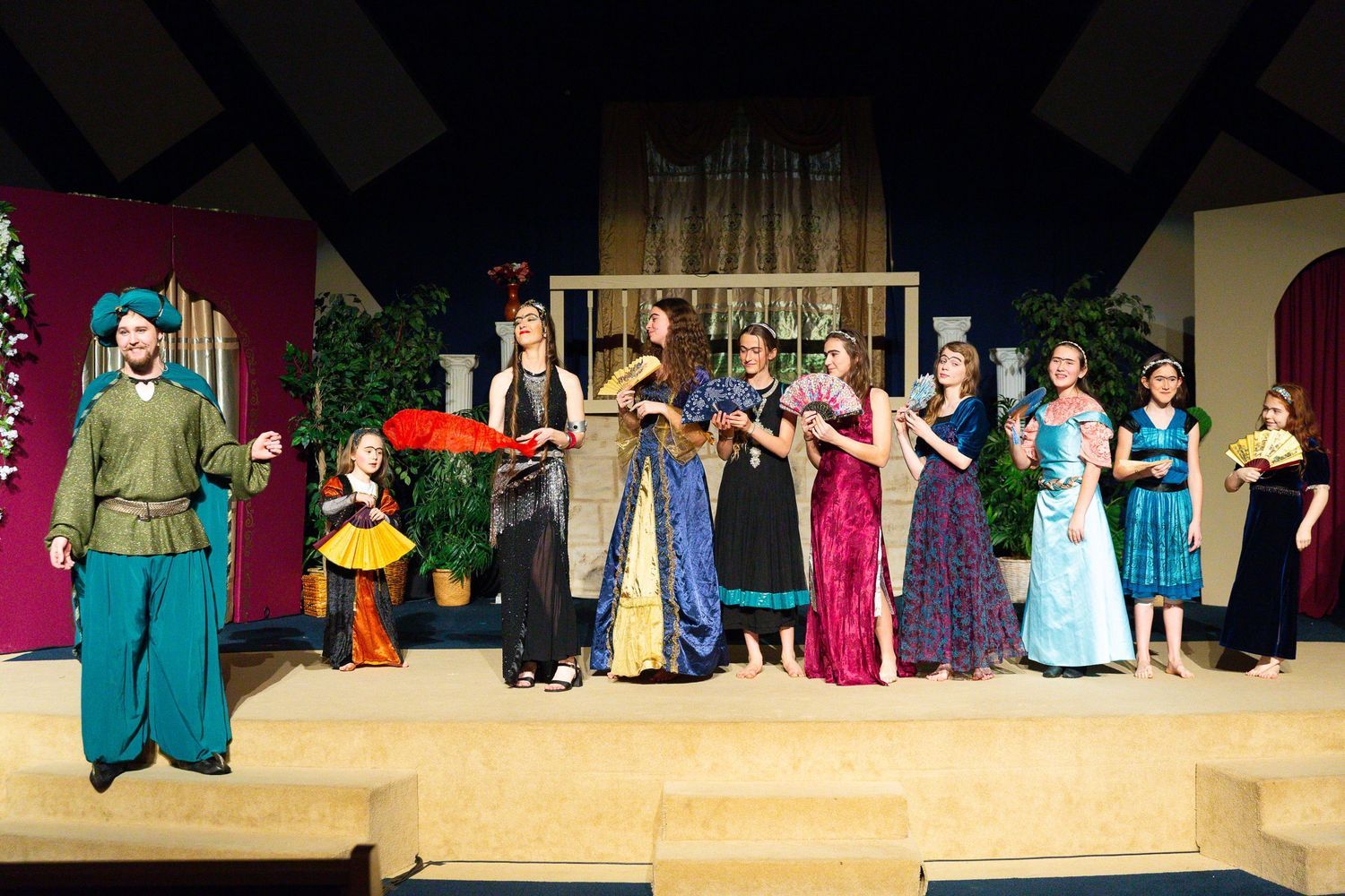 A cast of performers in colorful biblical-era costumes stands on a wooden stage set with architectural elements, plants, and draped fabrics, arranged in a formal group tableau.