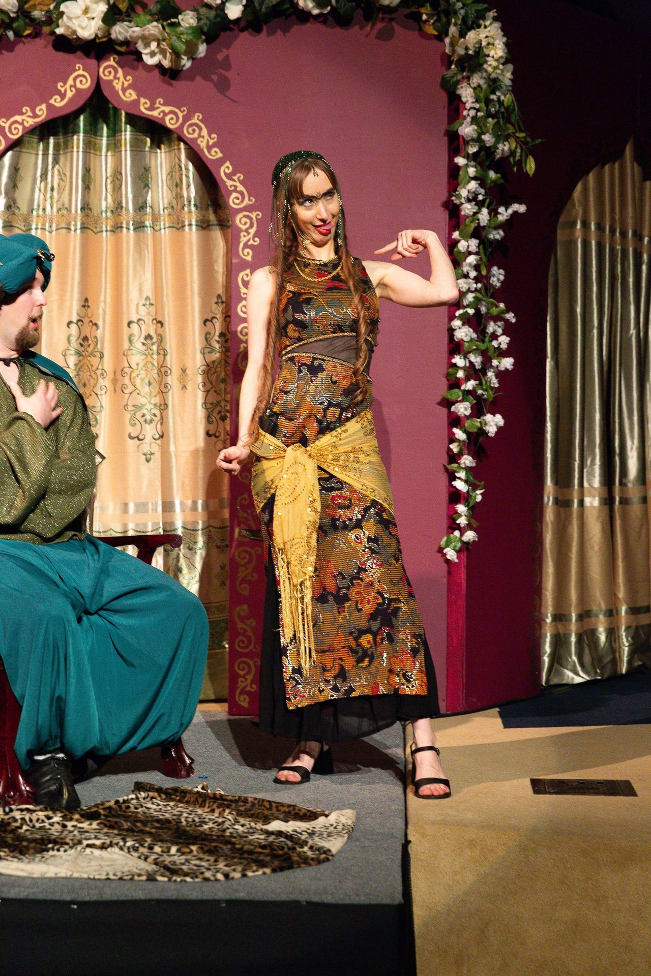 Two performers in ornate patterned costumes stand on stage before a mauve backdrop decorated with floral garland, with gold-trimmed curtains and decorative panels visible behind them.