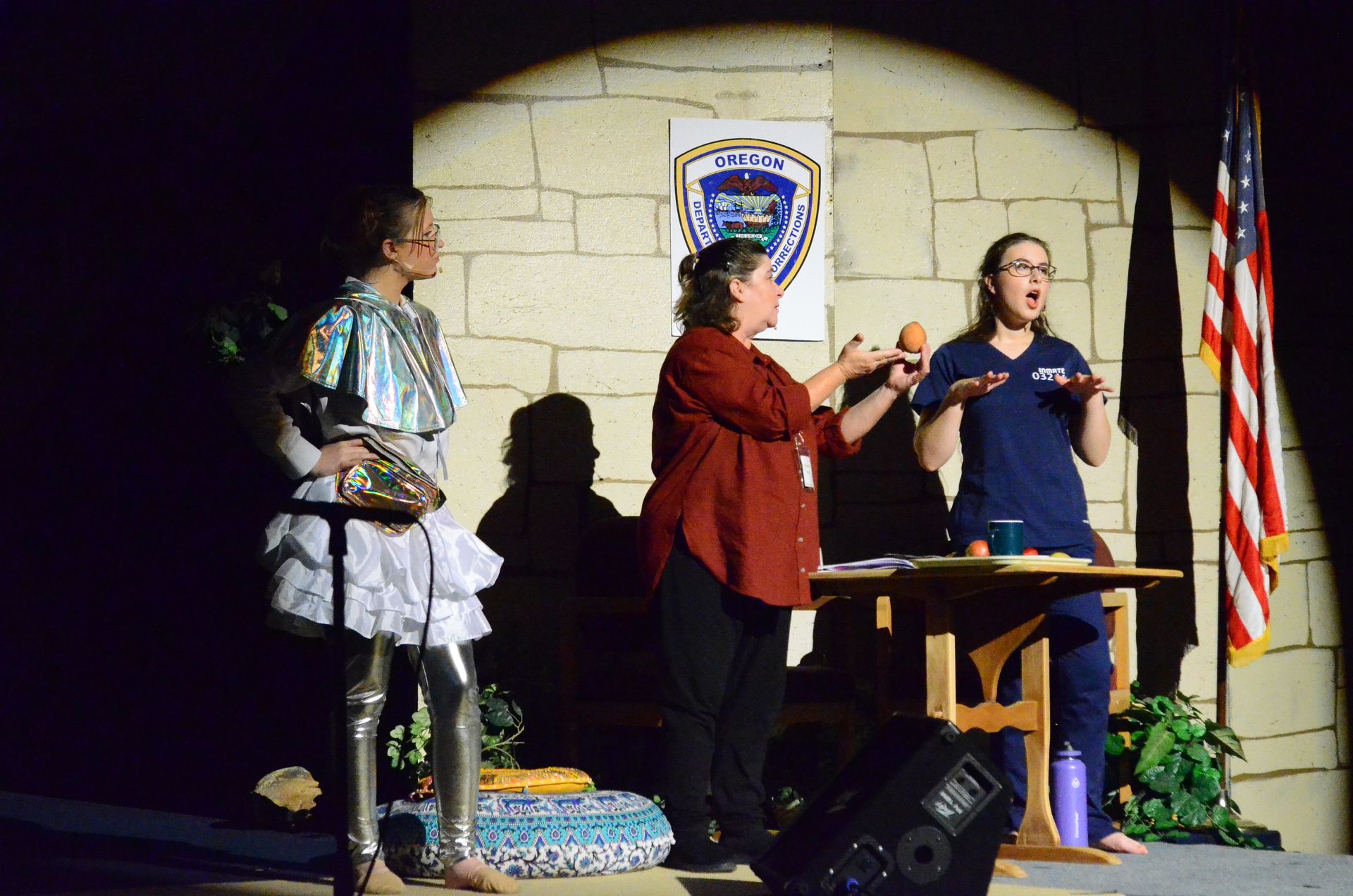 Three performers in colorful costumes stand on stage beside a wooden table with props, with an American flag and an Oregon police badge banner visible on the stone wall behind them.