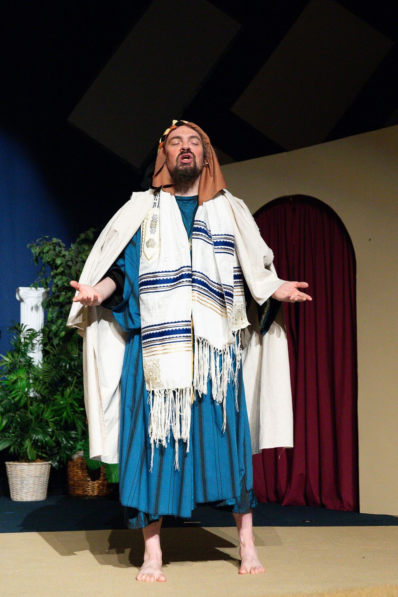 A performer stands barefoot on stage wearing a blue tunic, white robe, and decorative striped shawl, with arms outstretched in an expressive gesture, framed by potted plants and red curtains.