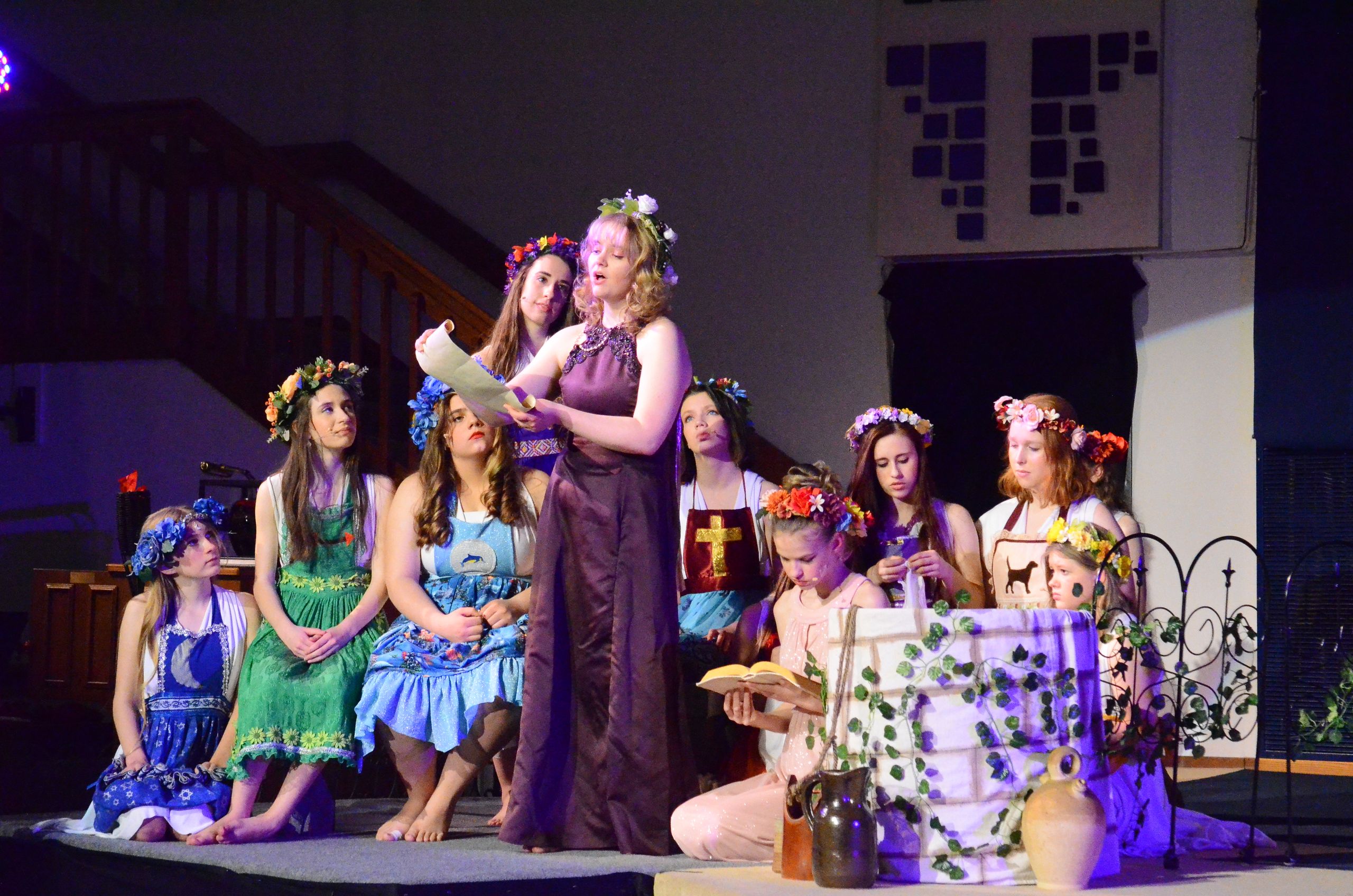 A cast of performers wearing colorful floral crowns and flowing costumes stands together on stage in a church setting, with one performer standing elevated holding a scroll while others gather around decorative props including pottery and greenery.