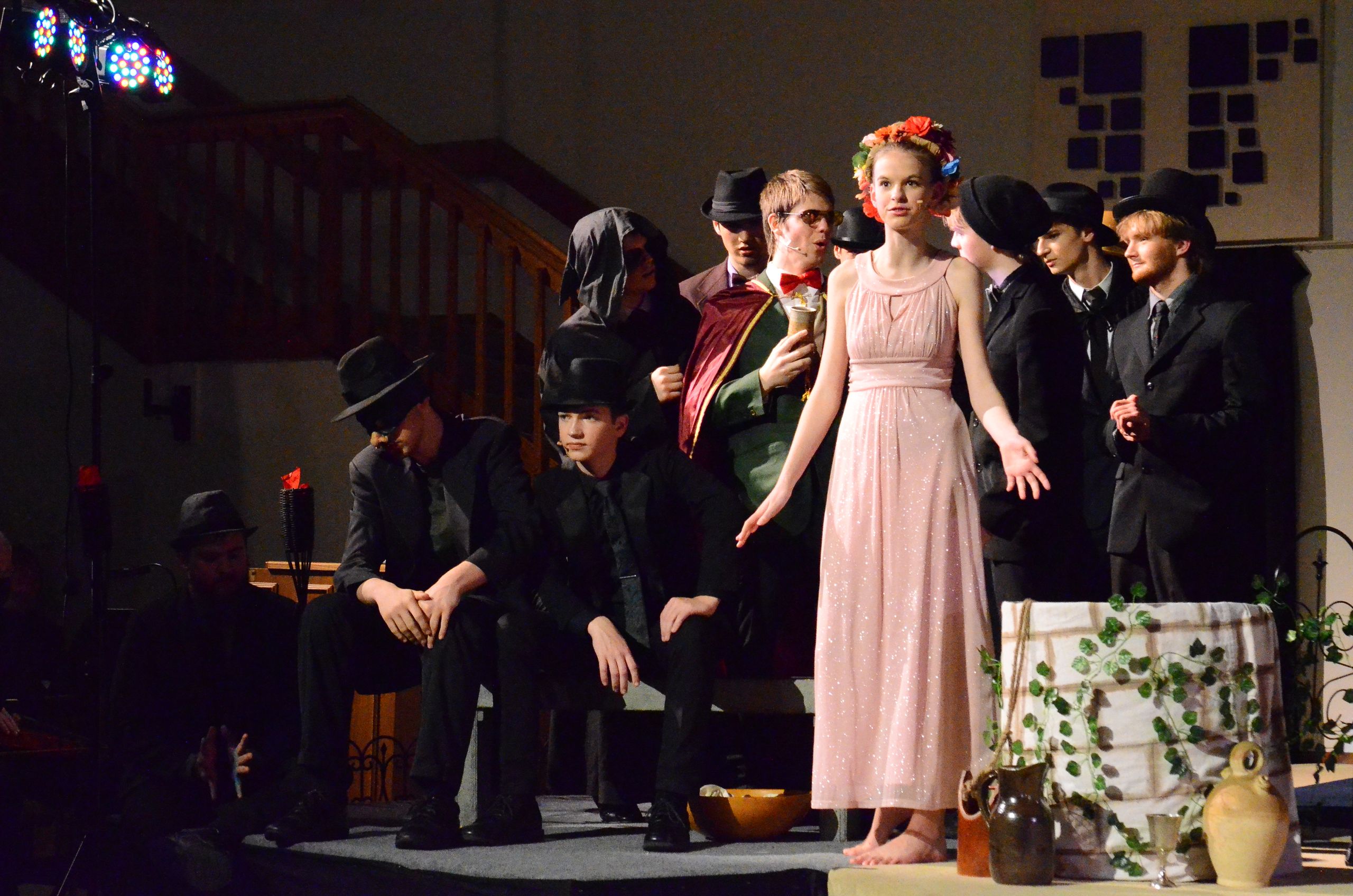 A cast of performers in period costumes stands on stage, with some seated wearing dark suits and fedoras on the left, while others stand in the background, and a central performer in a pink dress with a flower crown stands prominently under colorful stage lighting, surrounded by props including plants and pottery.