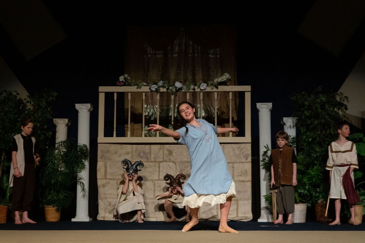 A performer in a light blue nightgown with lace trim stands center stage with arms outstretched in joy, surrounded by other cast members in period costumes on a set featuring classical columns, a wooden structure, potted plants, and decorative architectural elements.
