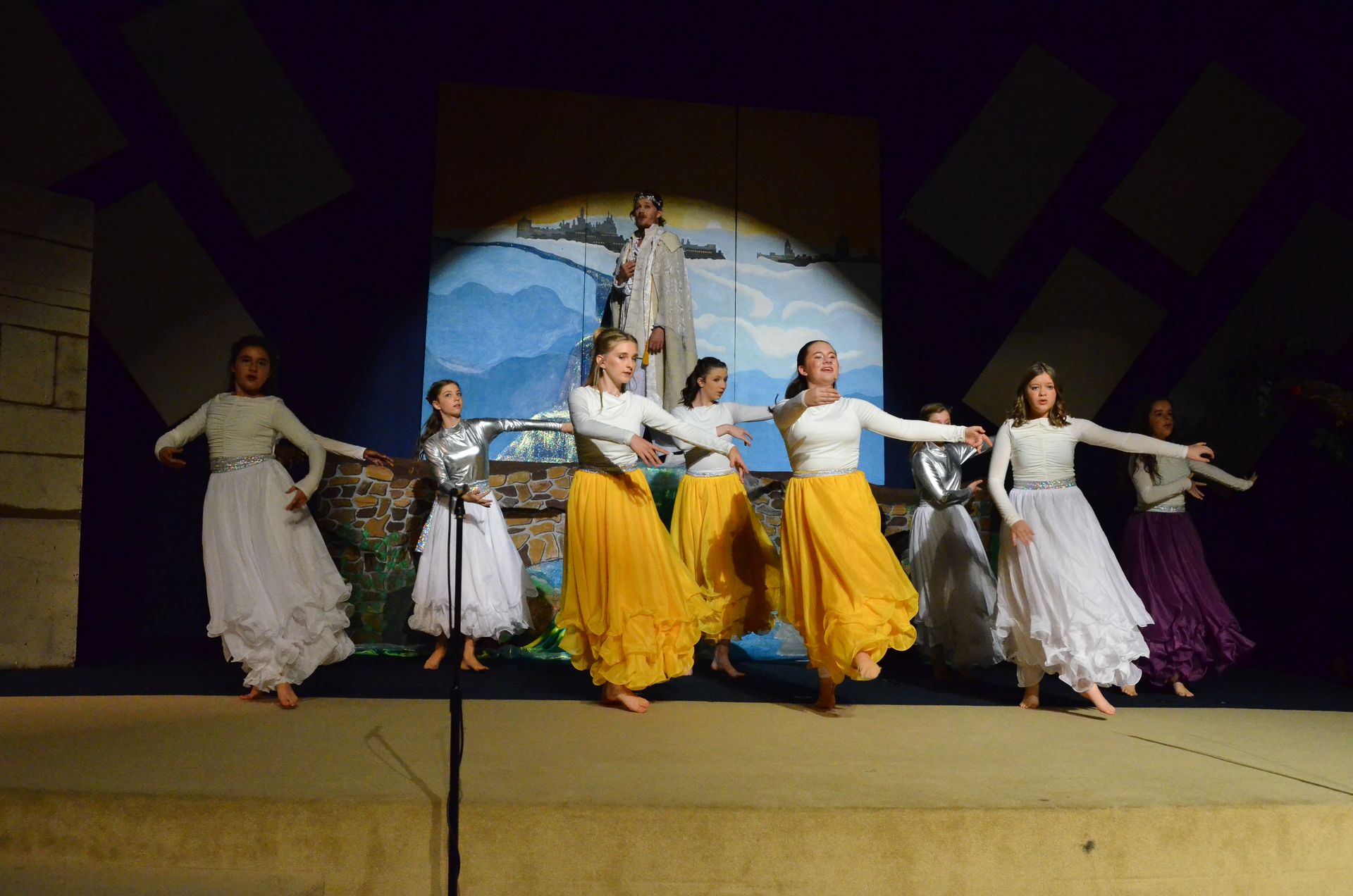 A line of performers in white and yellow flowing skirts and white tops stand barefoot on stage with arms outstretched, performing in front of a painted backdrop depicting mountains and a coastal landscape with a figure in robes.