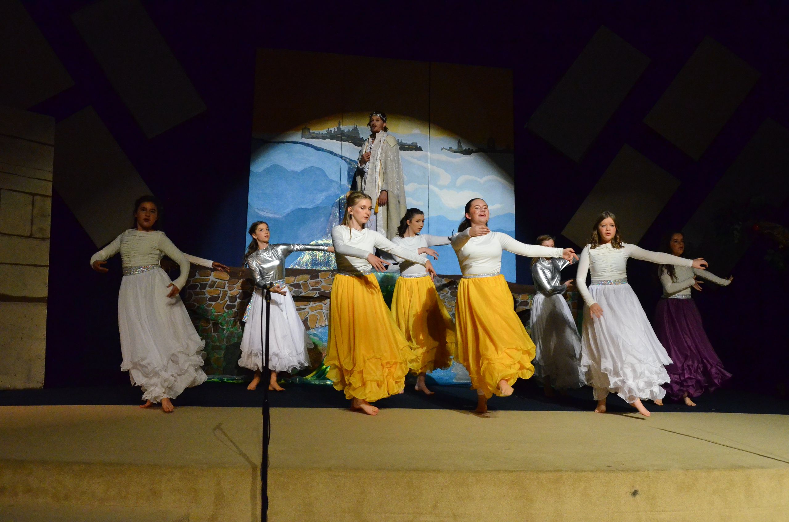 A line of performers in white and yellow flowing skirts and white tops stand barefoot on stage with arms outstretched, performing in front of a painted backdrop depicting mountains and a coastal landscape with a figure in robes.