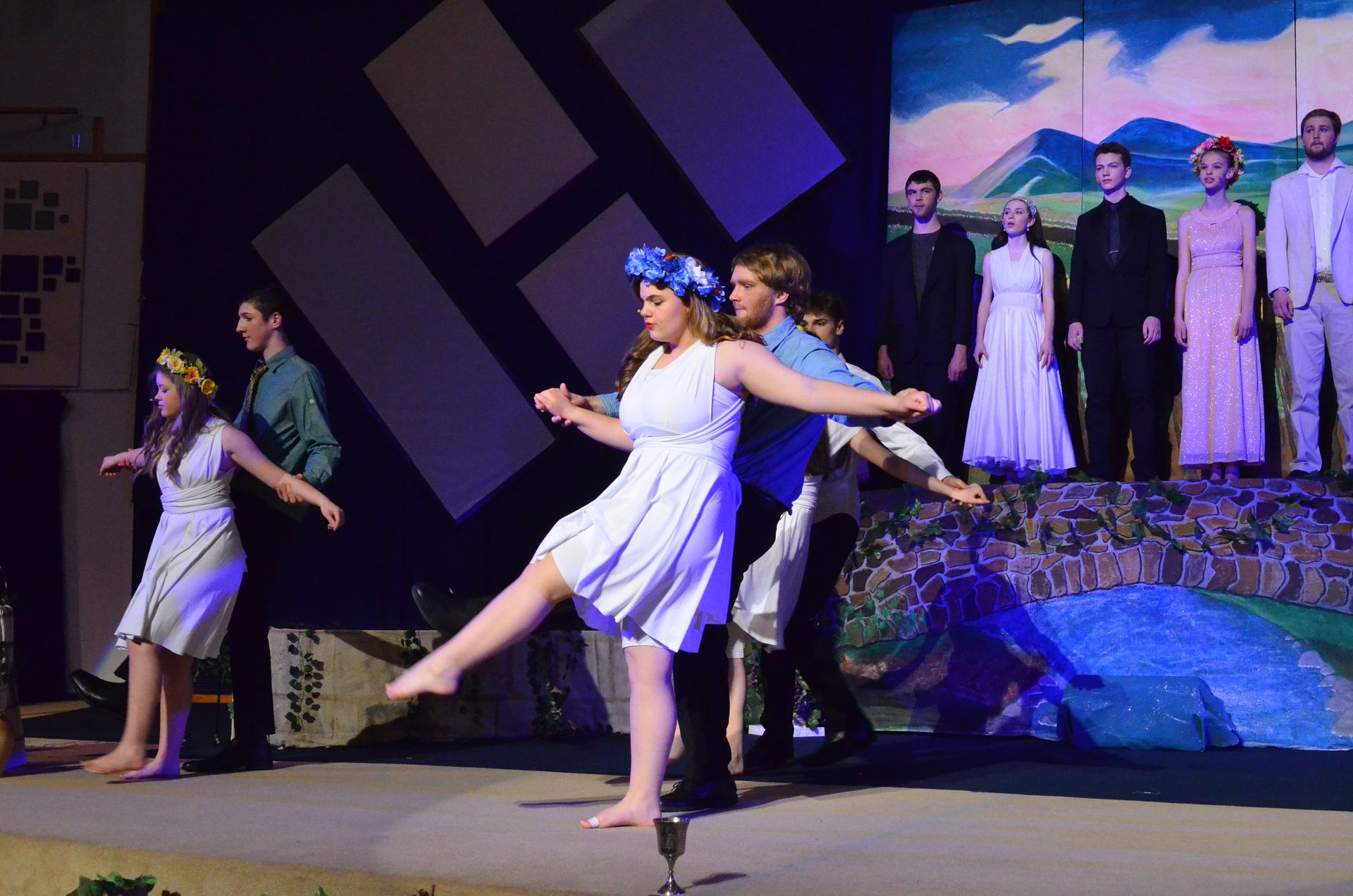 A performer in a white dress with a blue flower crown dances expressively center stage while other cast members stand in formation on either side, with abstract geometric backdrop elements and a painted landscape scene visible behind them.
