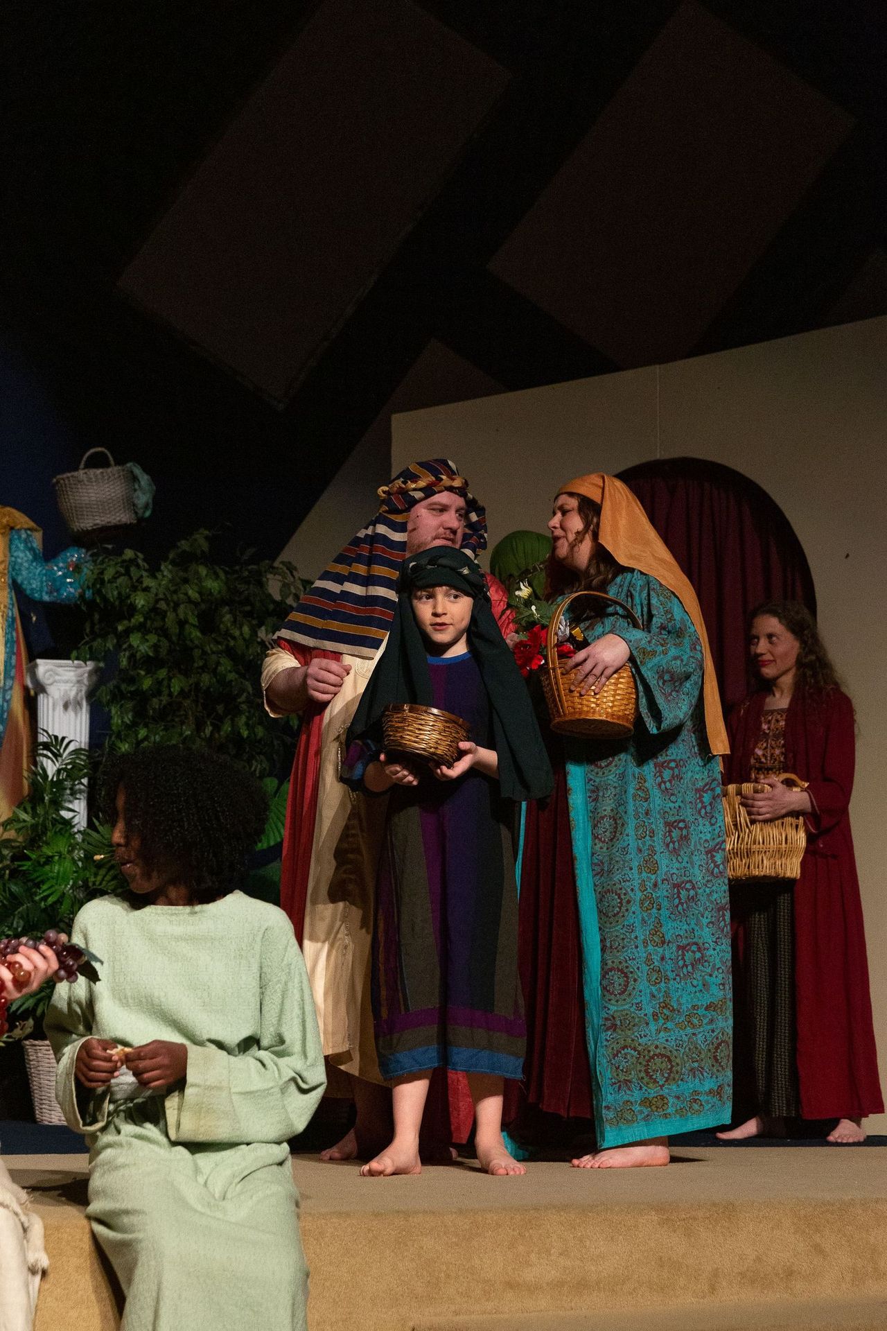 Cast members in colorful biblical-era costumes stand on stage holding woven baskets, with potted plants and draped fabric set pieces visible in the background.