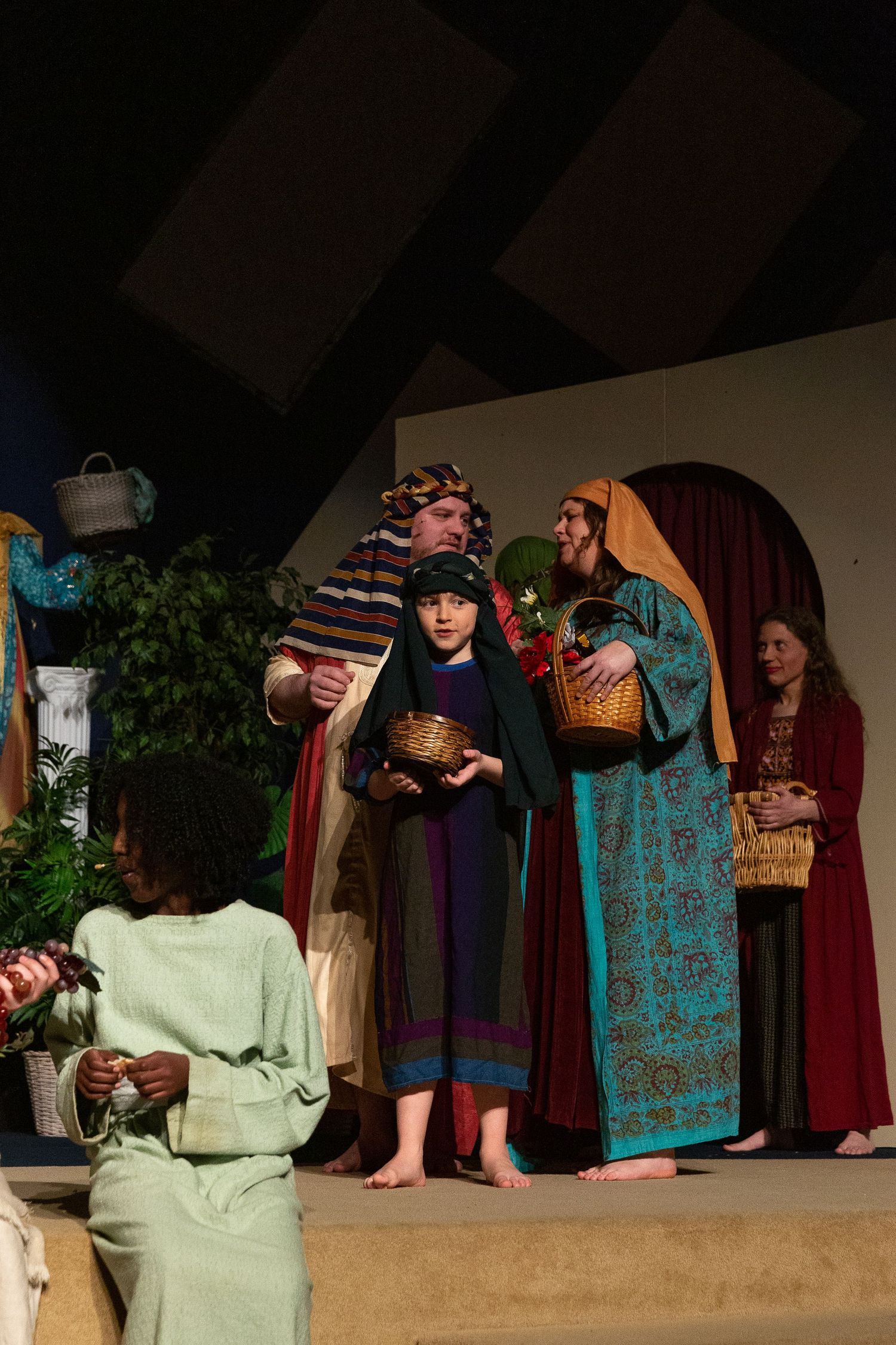 Cast members in colorful biblical-era costumes stand on stage holding woven baskets, with potted plants and draped fabric set pieces visible in the background.