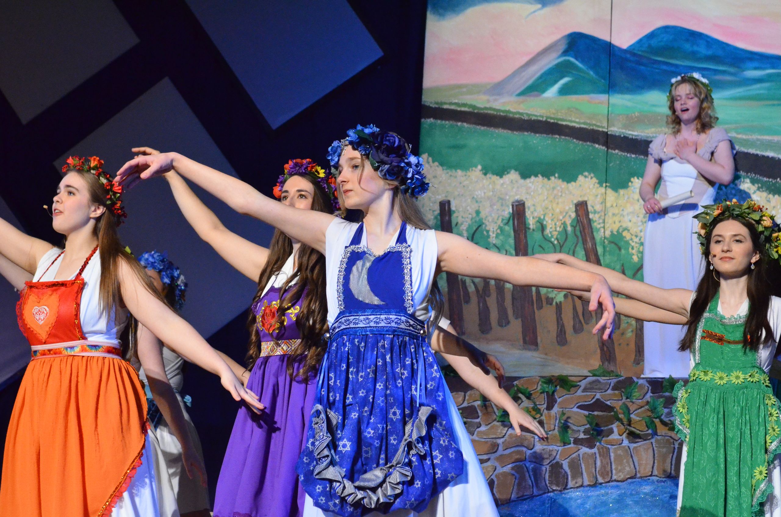 Four performers in colorful traditional folk dresses with flower crowns stand on stage in choreographed poses, with a painted backdrop depicting mountains and fields behind them.