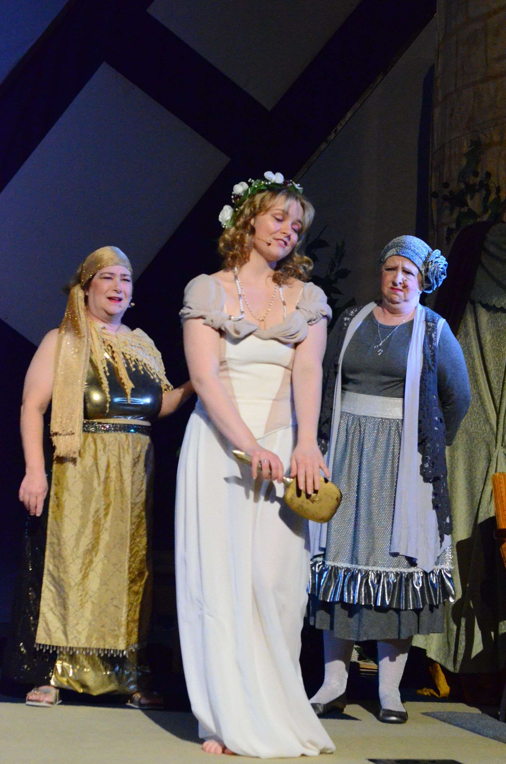 Three performers in costume stand on an indoor stage, with one wearing a blue headwrap and striped shawl, another in a cream-colored dress with floral headpiece holding a yellow object, and a third in a shimmering gold garment.