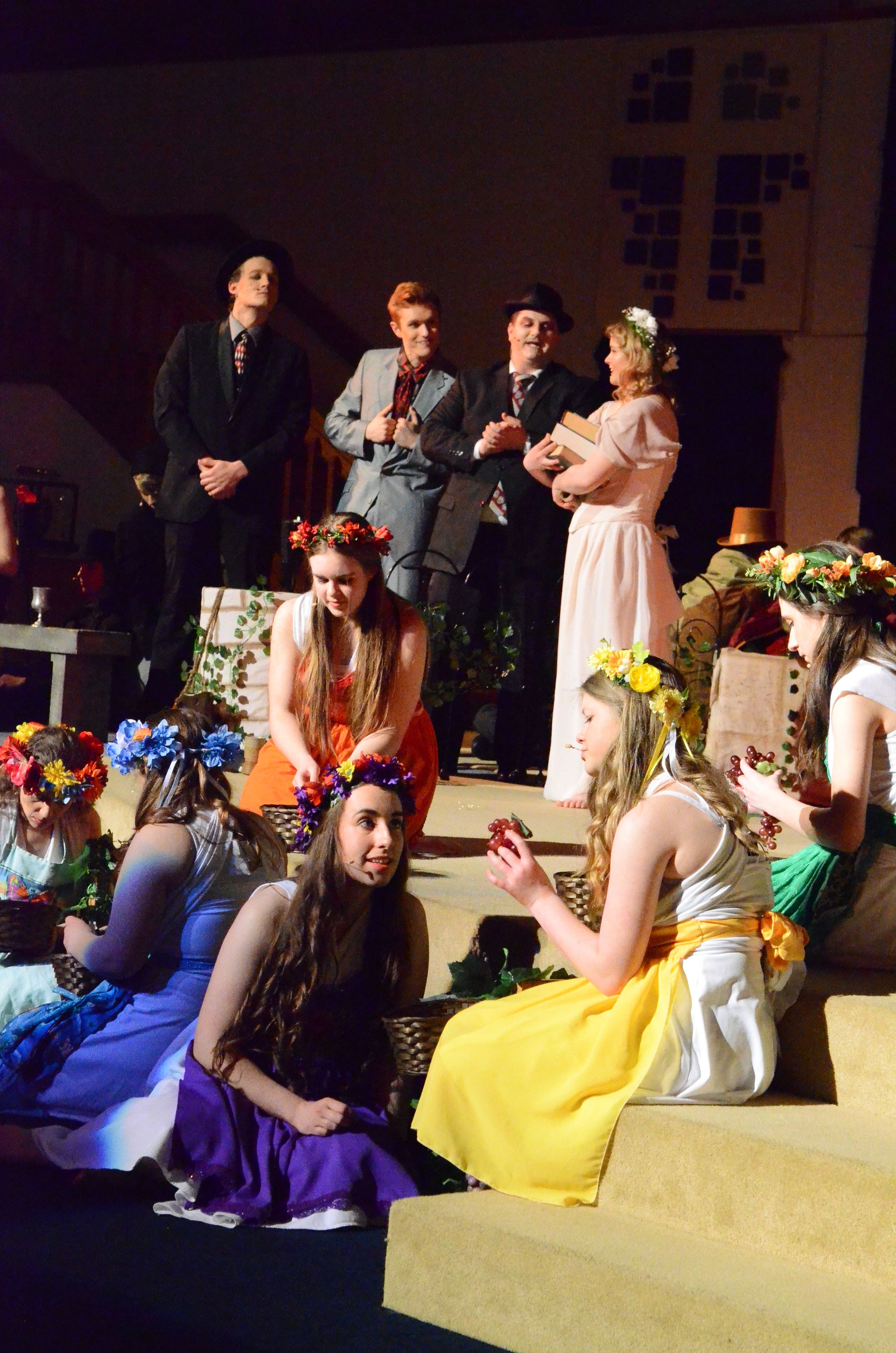 A cast of performers in colorful costumes adorned with floral garlands stands together in an indoor venue, arranged in a theatrical pose for a musical production.