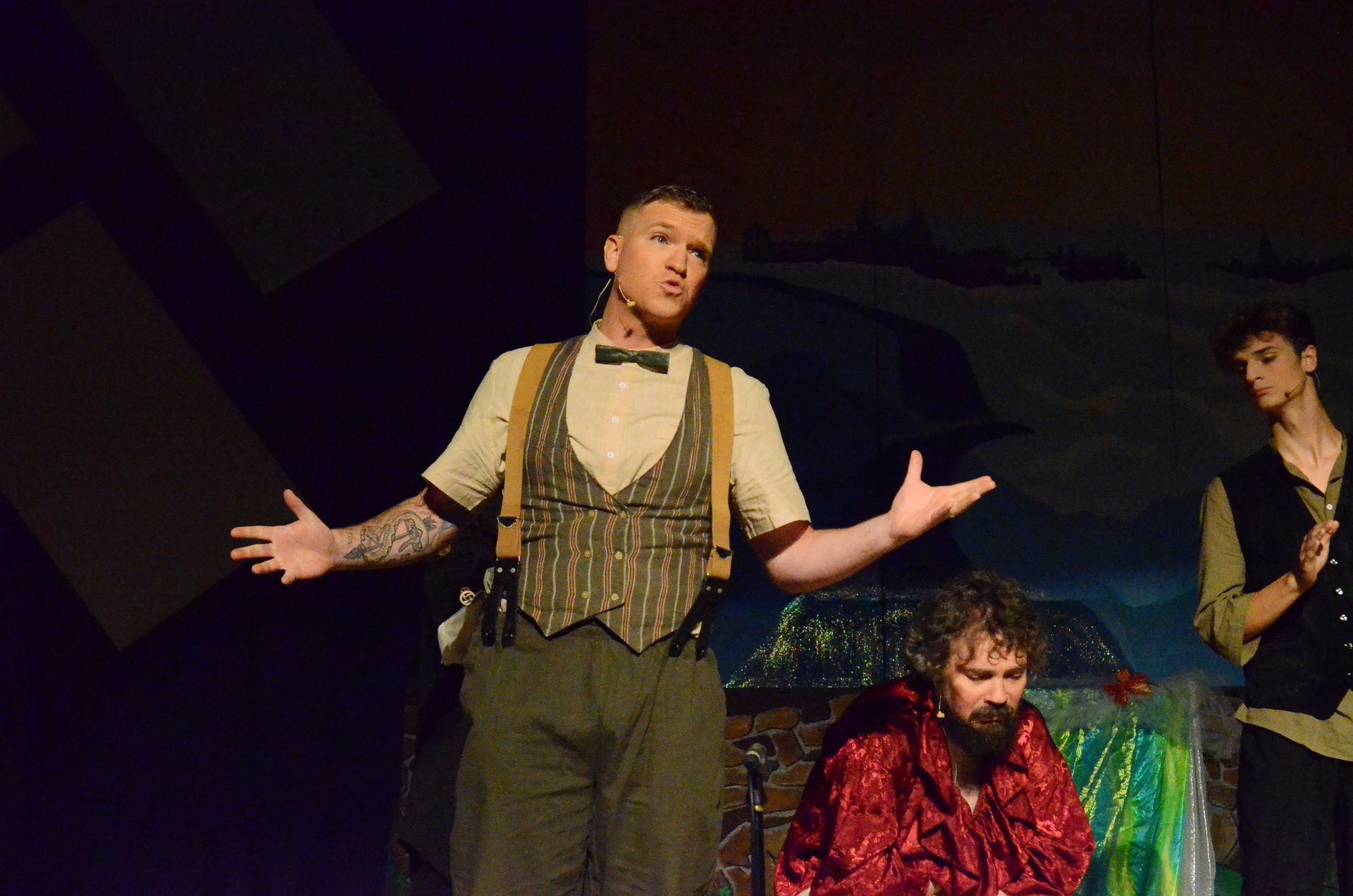 A performer in a striped vest and suspenders gestures expressively center stage while another performer in a red velvet jacket sits nearby, with additional cast members visible in the dimly lit theatrical setting.