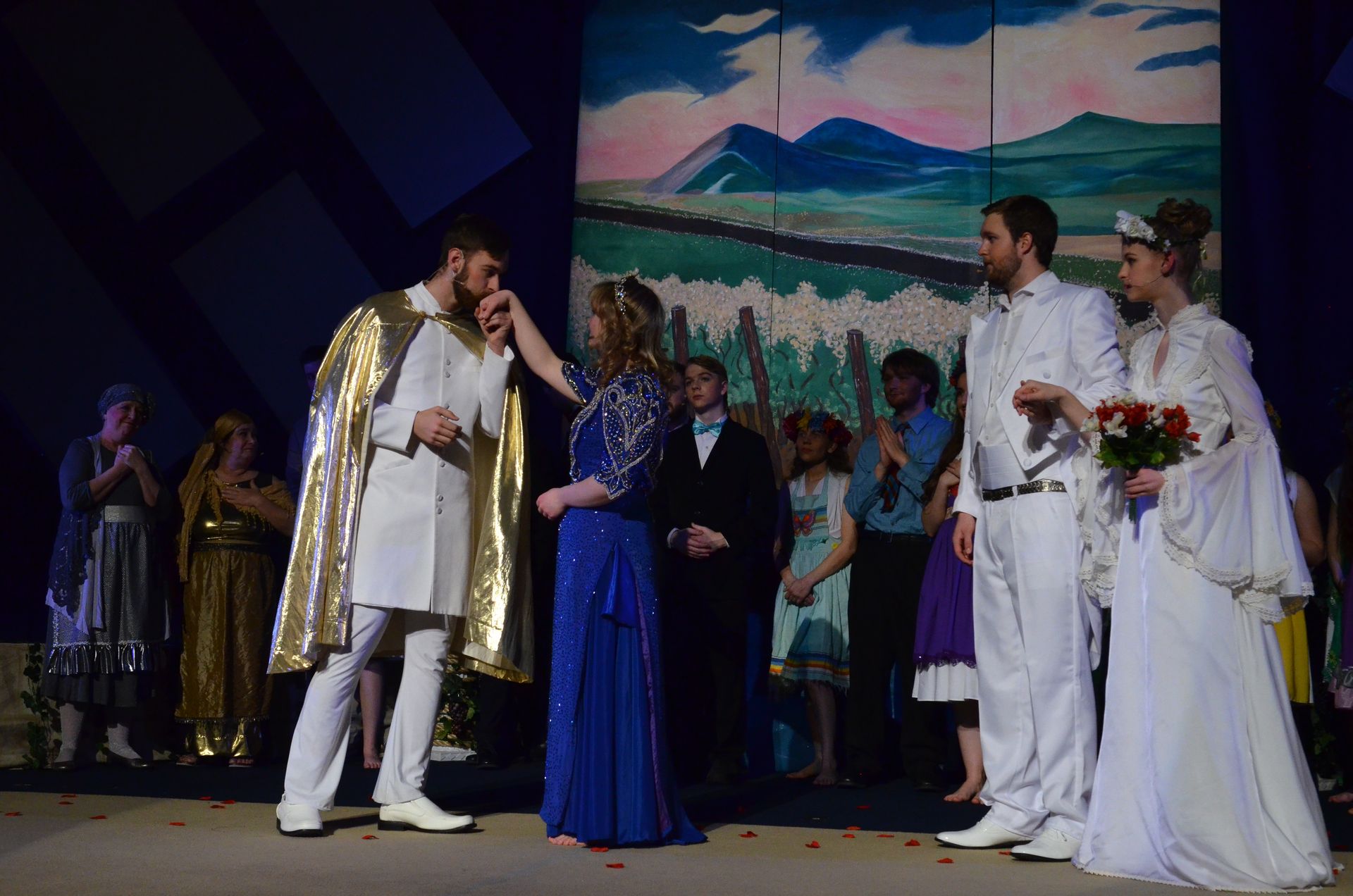 A cast of performers in white and gold robes and blue period costumes stands on stage before a painted landscape backdrop, with the central performer in a gold-trimmed white robe gesturing expressively while others watch, and scattered rose petals on the stage floor.