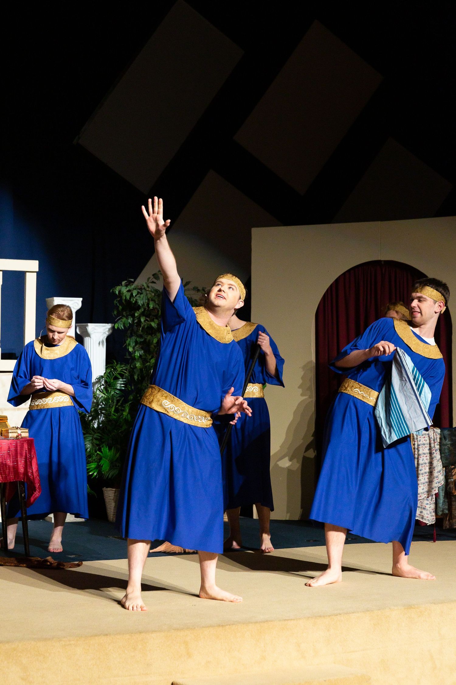 Performers in blue robes with gold trim stand barefoot on stage, with one raising their hand upward, surrounded by set pieces including a potted plant and wooden structures, in what appears to be a biblical-themed theatrical production.