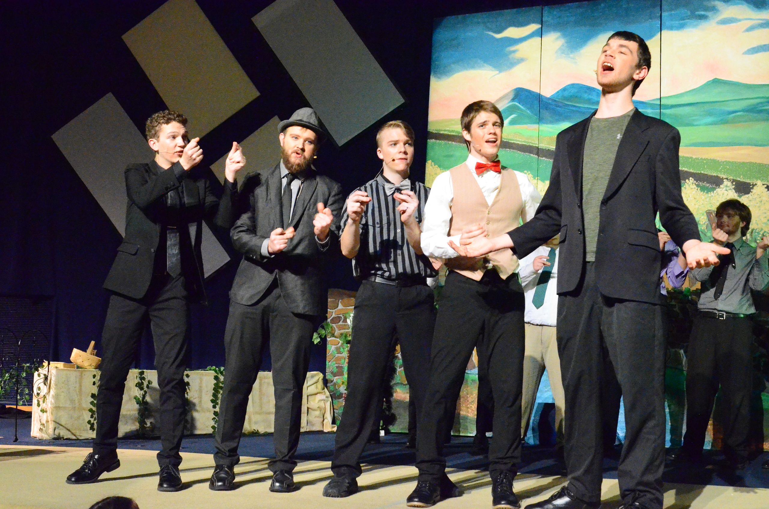 Five performers in formal black suits and vests stand on stage in front of a painted pastoral backdrop, gesturing expressively as they appear to be singing or performing together.