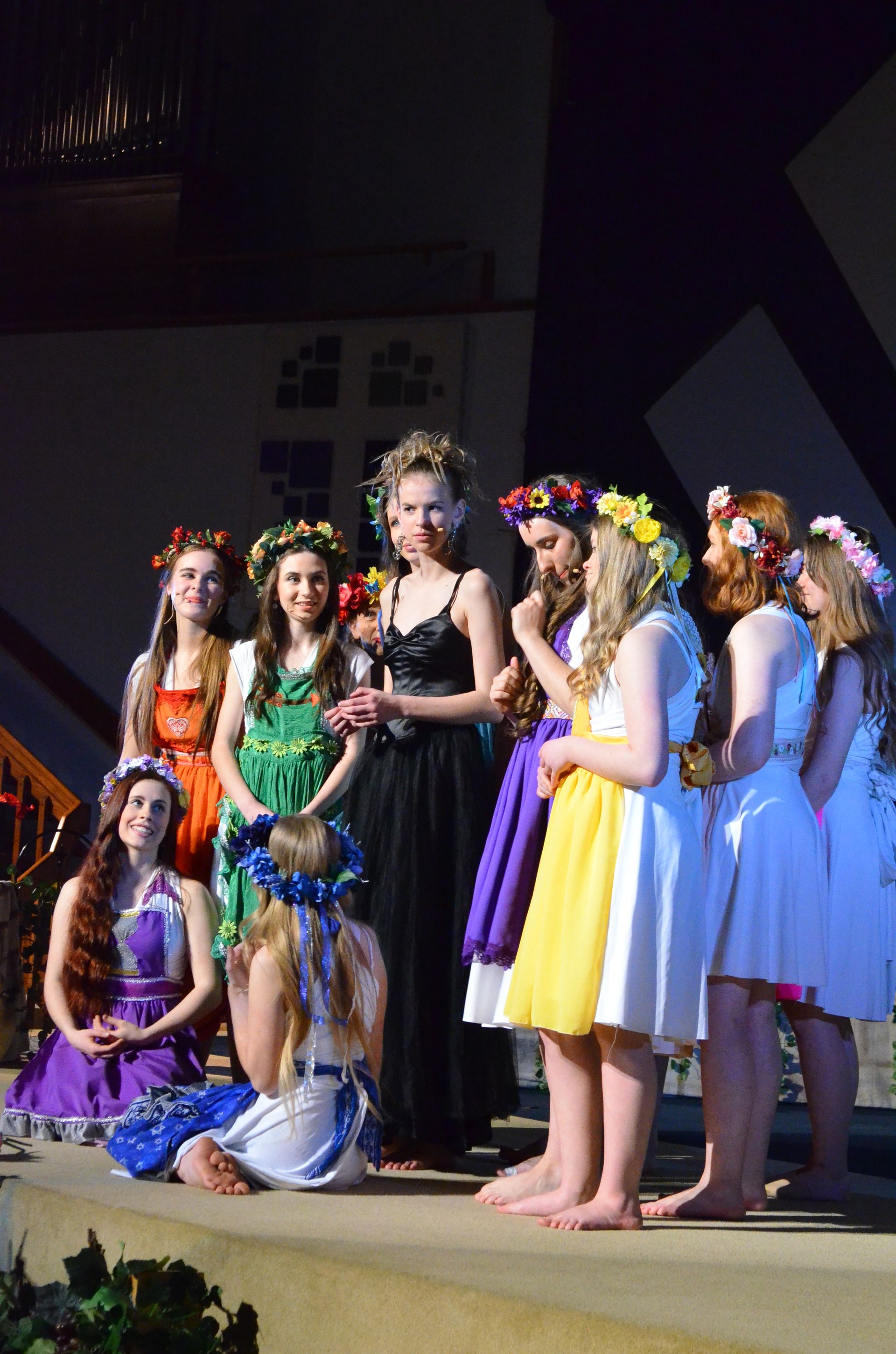 A cast of performers wearing colorful costumes and flower crowns stands in a line on an indoor stage, posed for a group photograph.