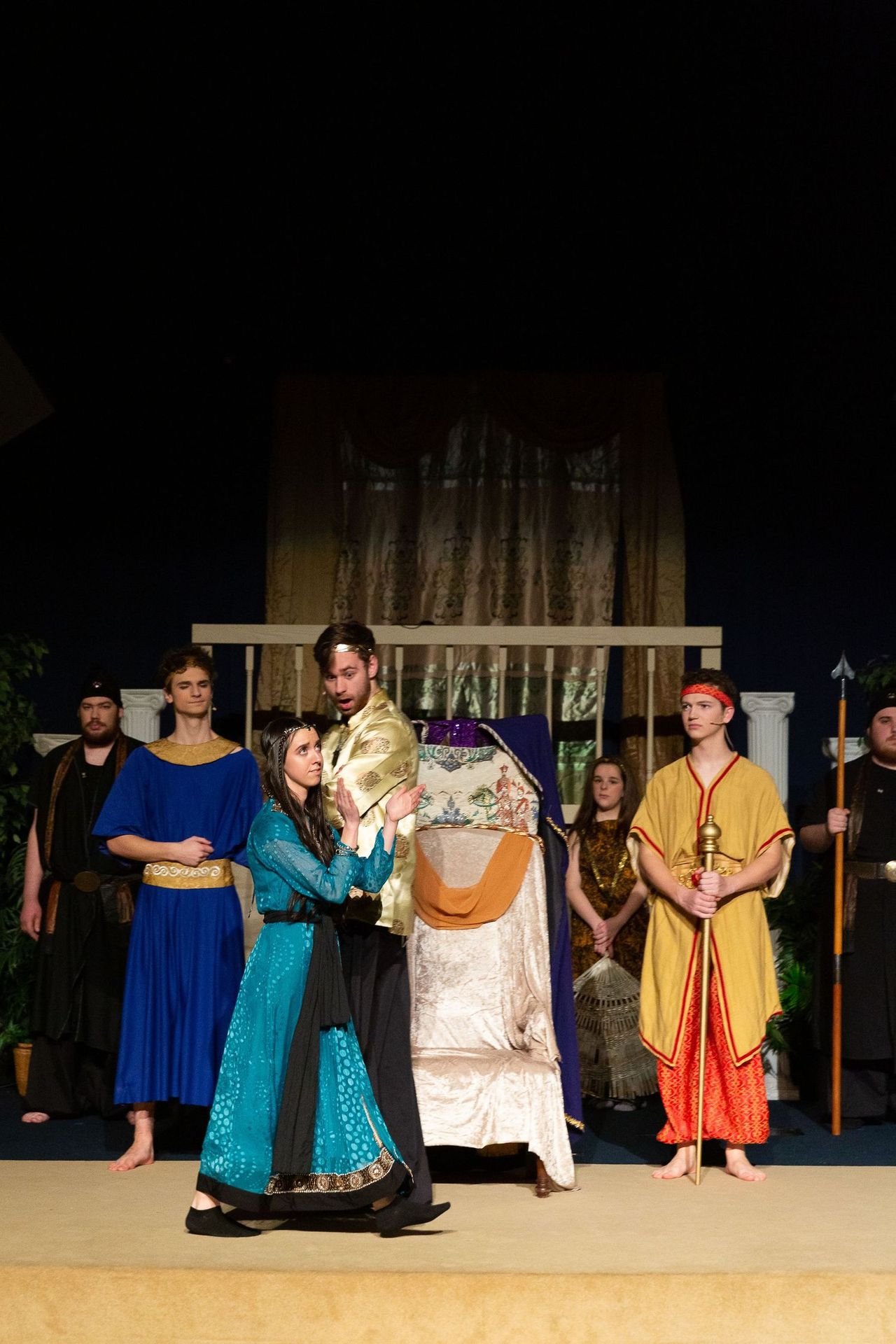 A cast of performers in colorful period costumes stands on stage before an ornate backdrop, with the central figures appearing to participate in a formal ceremonial scene.