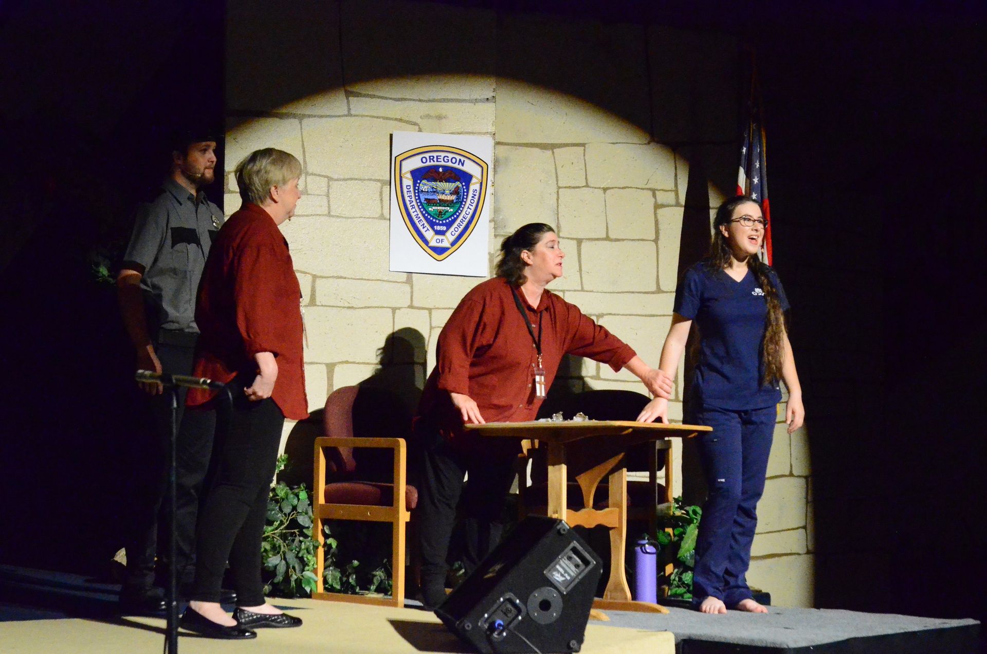A cast of performers stand on stage in period costumes before a stone wall backdrop featuring an Oregon law enforcement badge, with one performer seated at a wooden table and others positioned around the set.