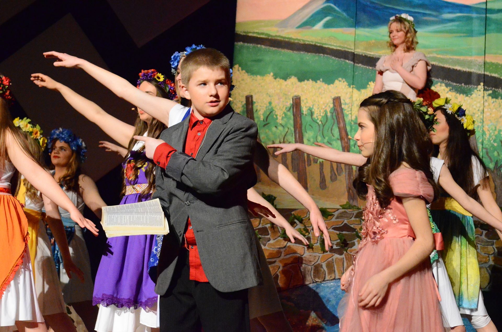 A young performer in a gray jacket and red shirt gestures expressively while surrounded by cast members wearing colorful costumes and flower crowns against a painted backdrop of a pastoral landscape.