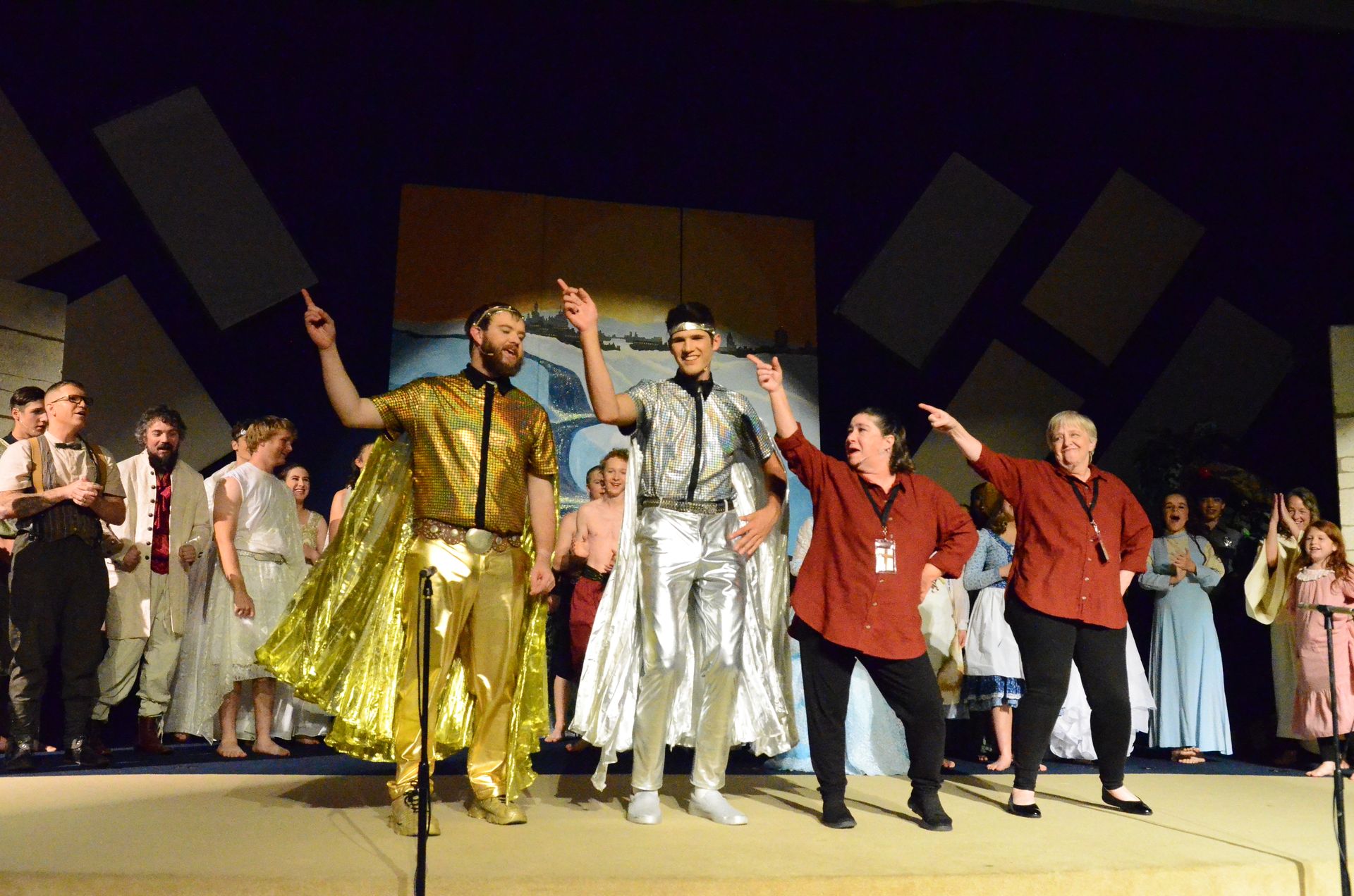 The cast takes a bow on stage in elaborate costumes including gold and silver lamé robes, with geometric set pieces and a landscape backdrop visible behind them.