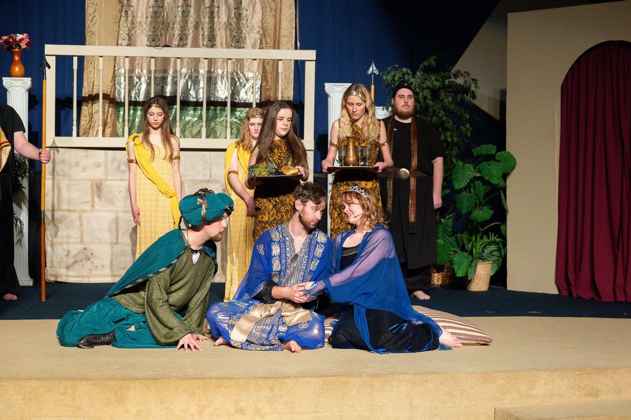 Cast members in ornate blue and gold robes perform a dramatic scene on stage, with performers kneeling and gesturing expressively while others in yellow and gold costumes stand in the background beside a set featuring classical columns, draped fabric, and potted plants.