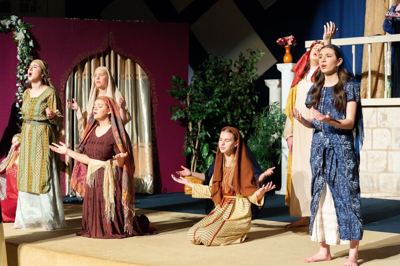 A cast of performers in flowing biblical-era costumes performs on a sand-covered stage set with ornate architectural elements, decorative plants, and draped fabrics in warm earth tones and jewel colors.