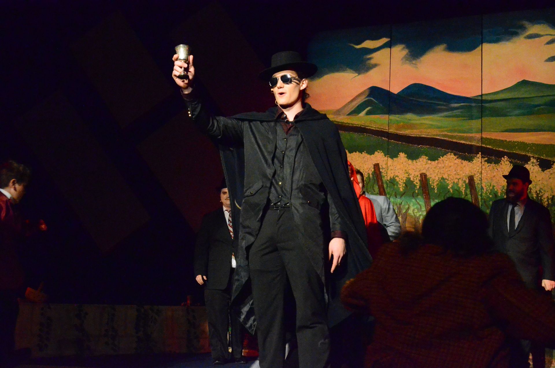 A performer in a black coat and top hat holds up a flashlight while standing on stage before a painted backdrop of a rural landscape at sunset, with other cast members visible in the scene.