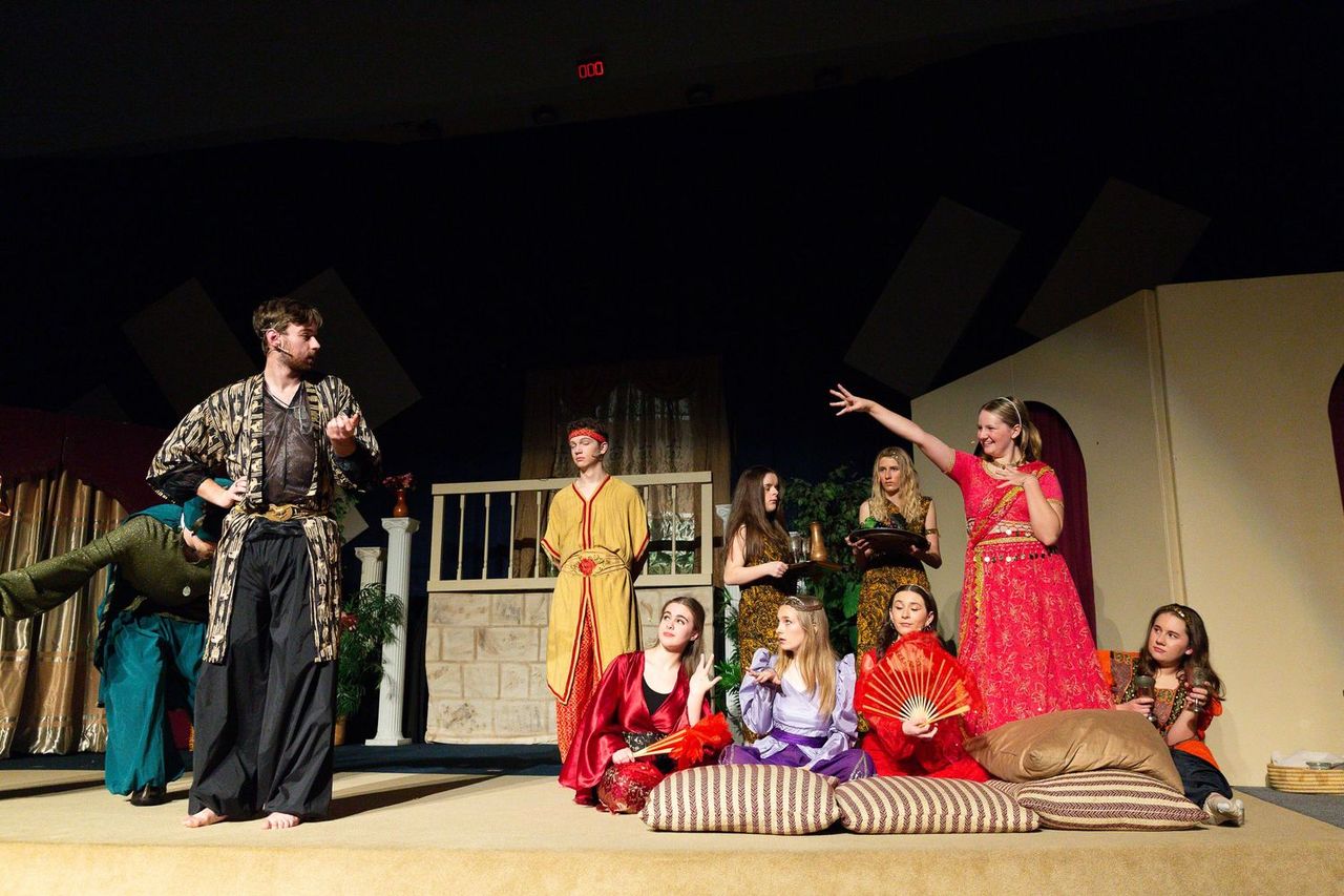 A diverse cast of performers in colorful robes and period costumes gathers on a warmly lit stage set with wooden furniture and potted plants, with some seated on striped cushions and others standing in dynamic poses.