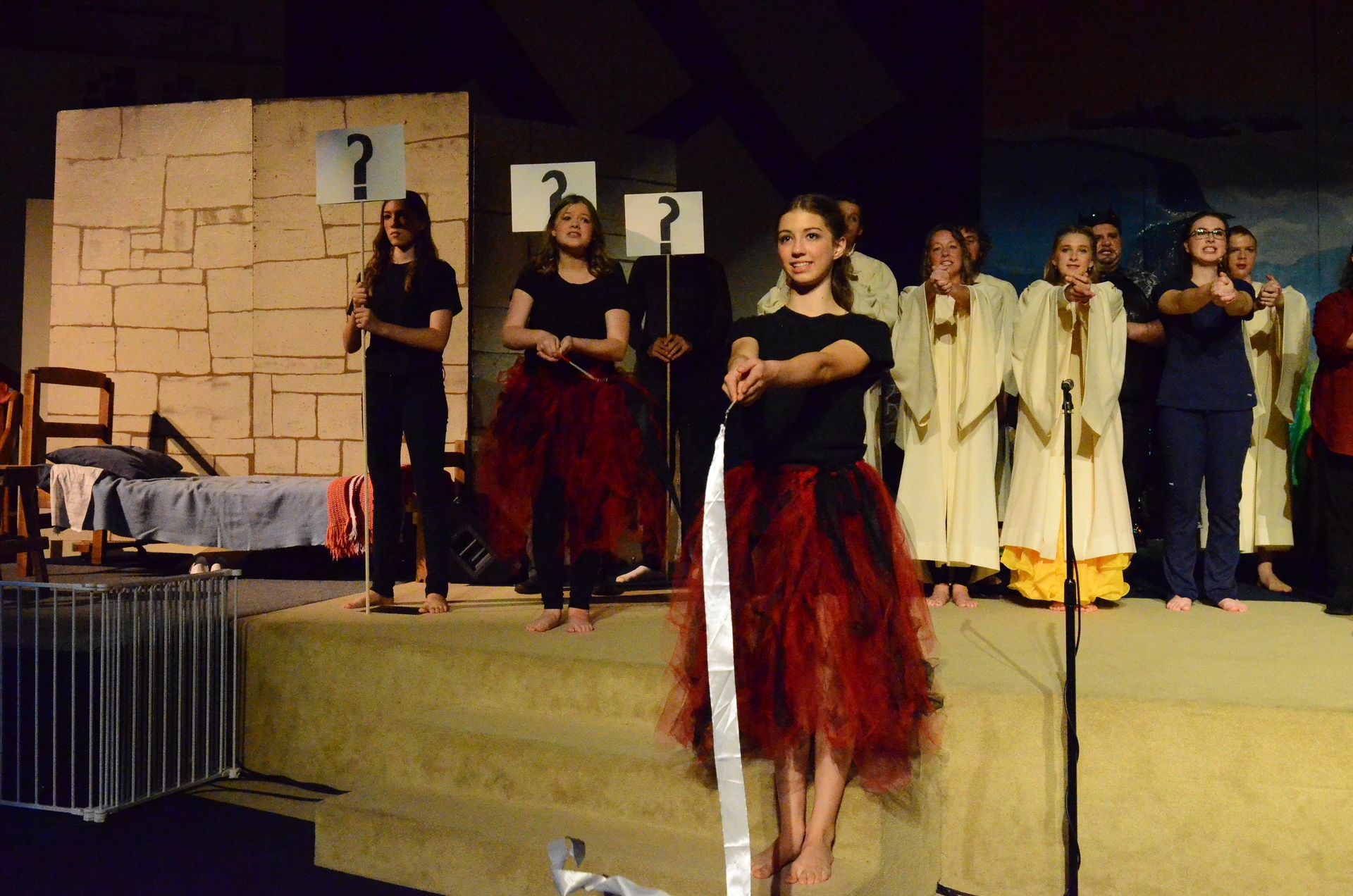 A cast of performers stands on stage in period costumes including red tunics and cream-colored robes, with some holding question mark signs, while a lead performer in a red dress sings at a microphone in front of a wooden block set piece.
