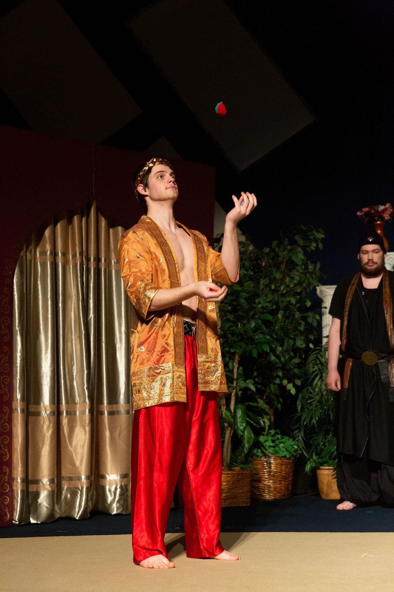 A performer in a gold and red costume jugles a red ball while looking upward on stage, with another costumed performer visible to the right and draped curtains and potted plants in the theatrical set behind them.