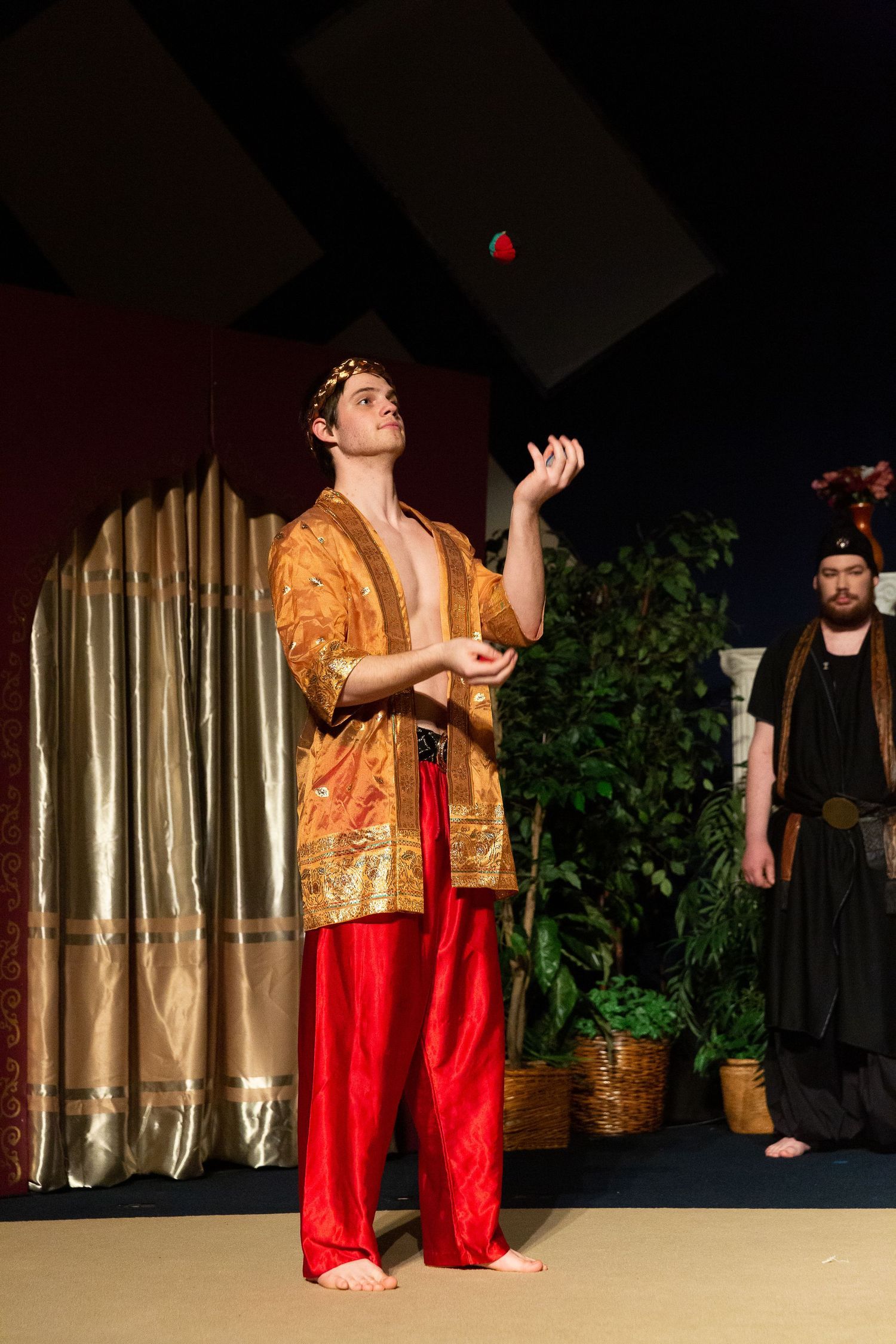 A performer in a gold and red costume jugles a red ball while looking upward on stage, with another costumed performer visible to the right and draped curtains and potted plants in the theatrical set behind them.