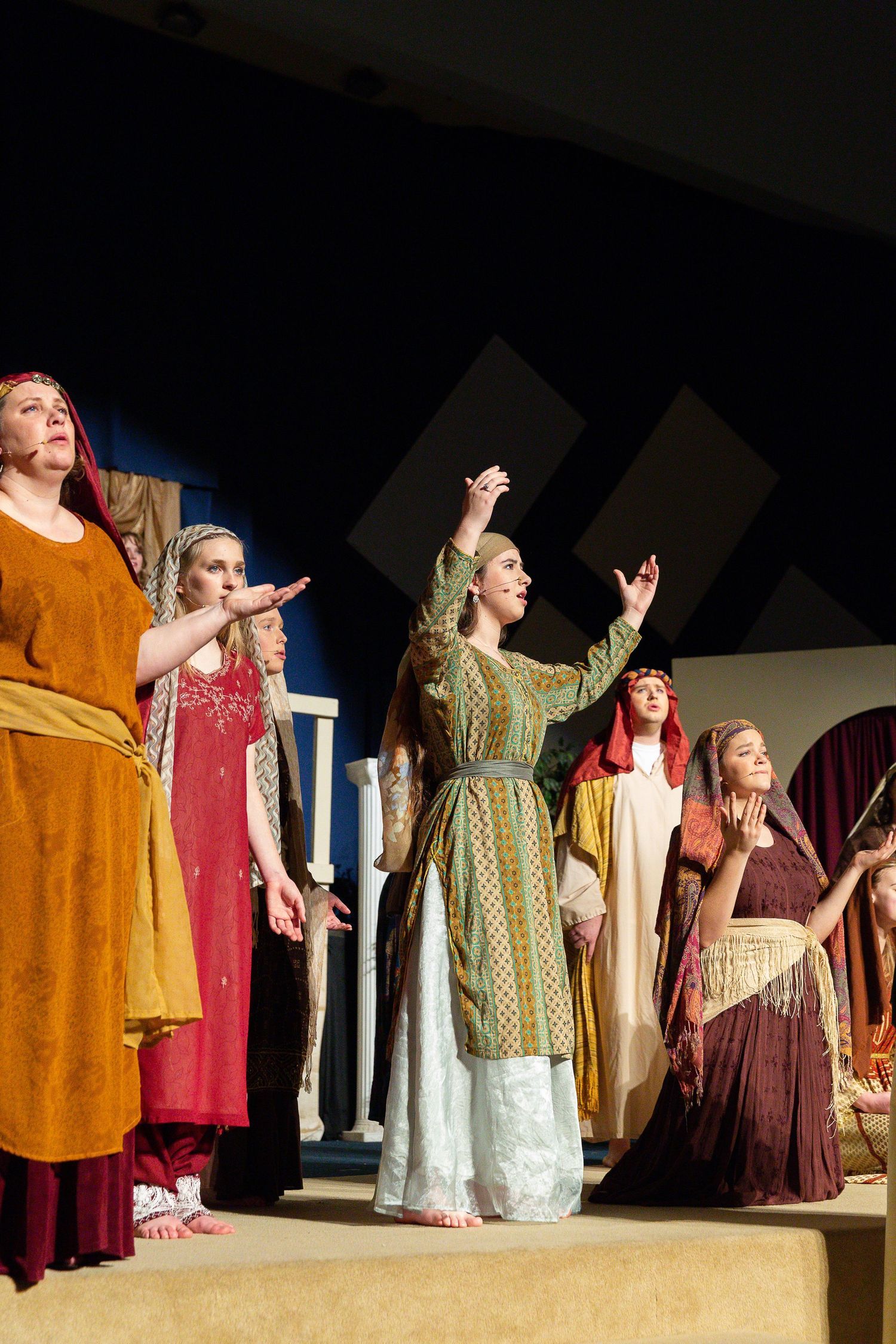 A cast of performers in richly colored biblical-era tunics and robes stands on stage with arms raised in gestures of wonder and supplication against a dark backdrop with geometric set pieces.