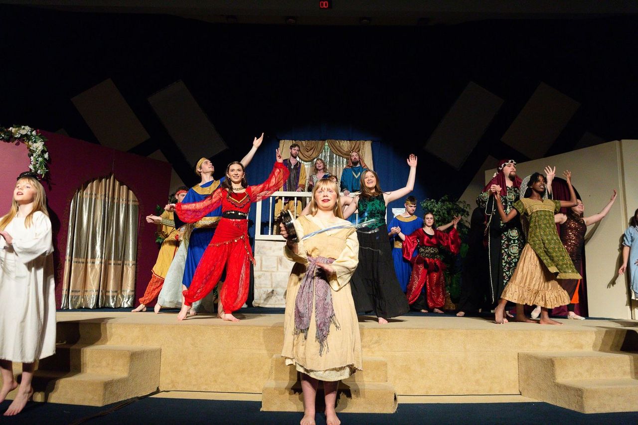 A large cast of performers in colorful biblical-era costumes gestures expressively on a multi-level stage set decorated with draped fabrics, potted plants, and an ornate backdrop, with one performer in a cream-colored robe and purple shawl standing prominently in the foreground.
