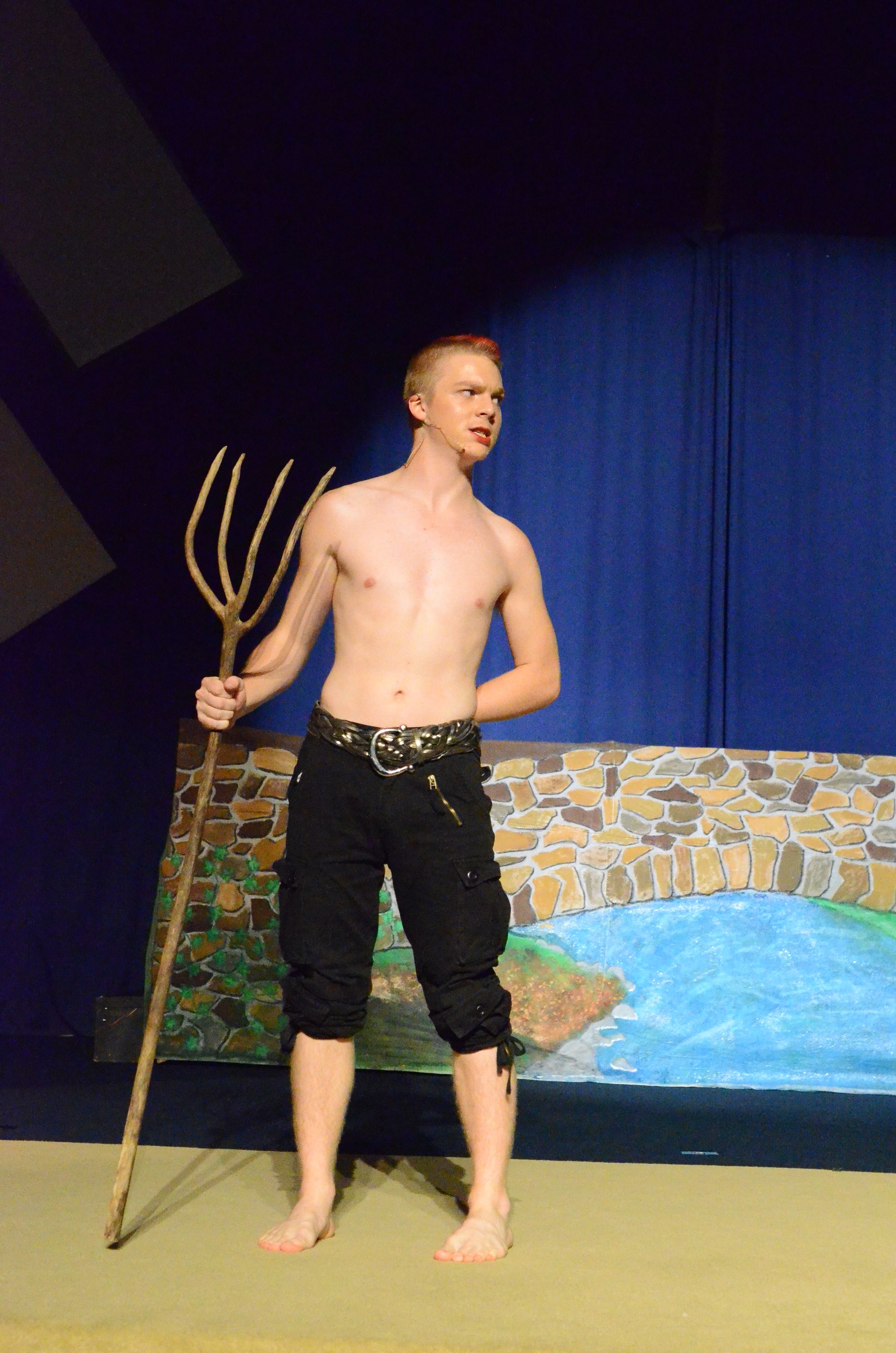 A shirtless performer in black pants and a gold chain belt reclines dramatically against a colorful painted backdrop depicting water and stone while holding a rope, against a blue wall in a theatre setting.
