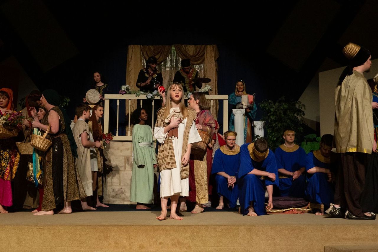 A cast of performers in biblical-era costumes stands on a sandy stage set with a wooden structure and draped fabric backdrop, with some performers seated in blue robes while others stand in various earth-toned garments around them.