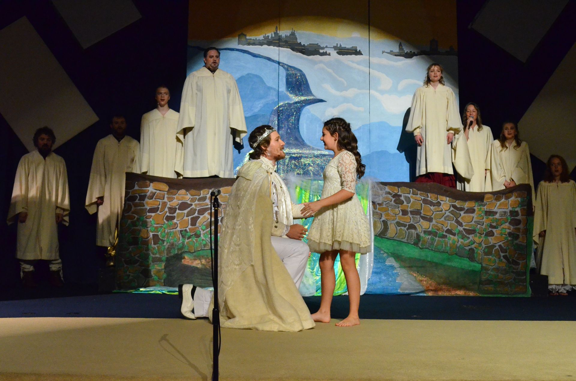 A cast of performers in cream-colored robes stands on stage around two central figures in conversation, with a backdrop depicting a coastal landscape with ships and a whale.