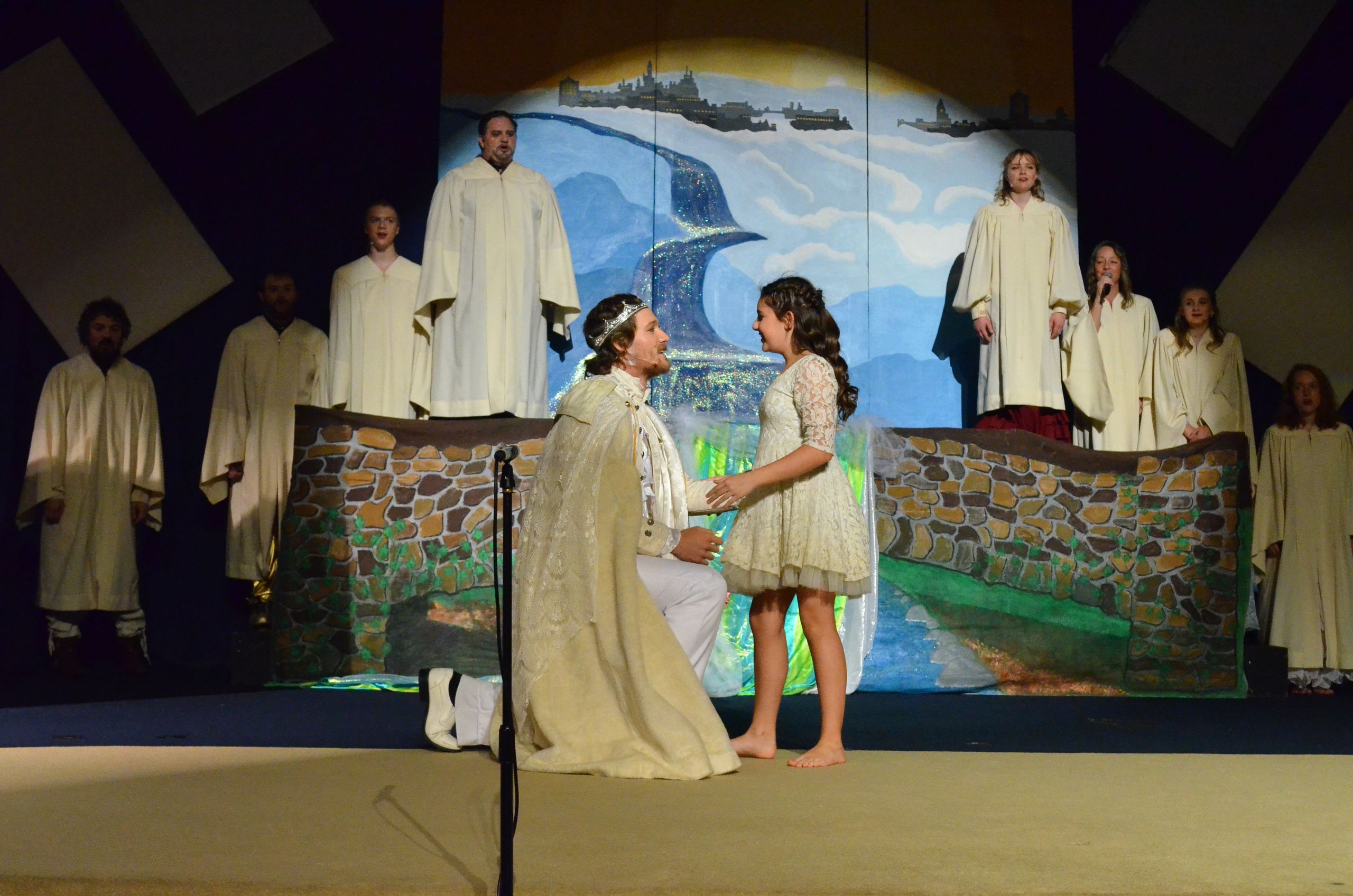 A cast of performers in cream-colored robes stands on stage around two central figures in conversation, with a backdrop depicting a coastal landscape with ships and a whale.