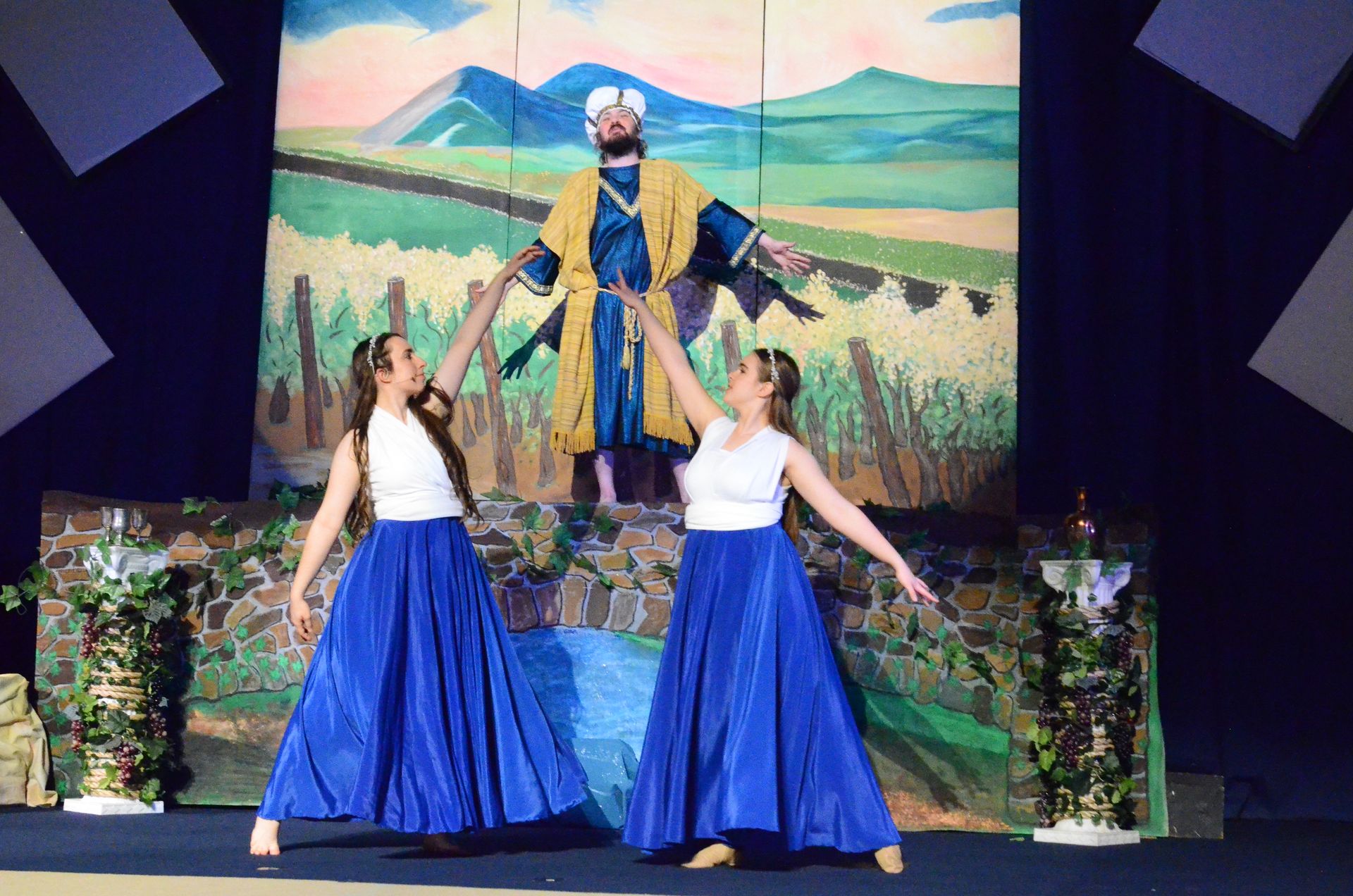 Two performers in white sleeveless tops and flowing blue skirts stand on stage before a large painted backdrop depicting rolling hills and mountains, with a giant figure in a yellow robe and white cap looming behind them against a stone wall set piece.