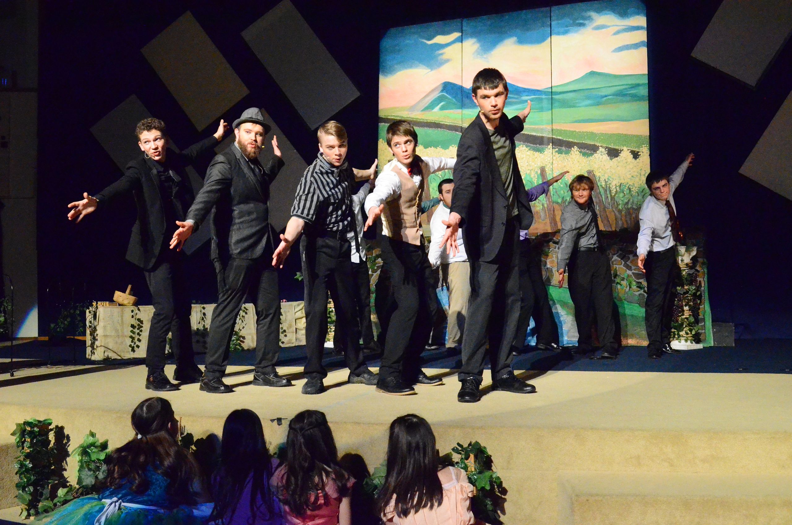 A cast of performers in dark suits and period costumes stands on stage in front of a painted backdrop depicting a mountainous landscape, with audience members visible in the foreground.