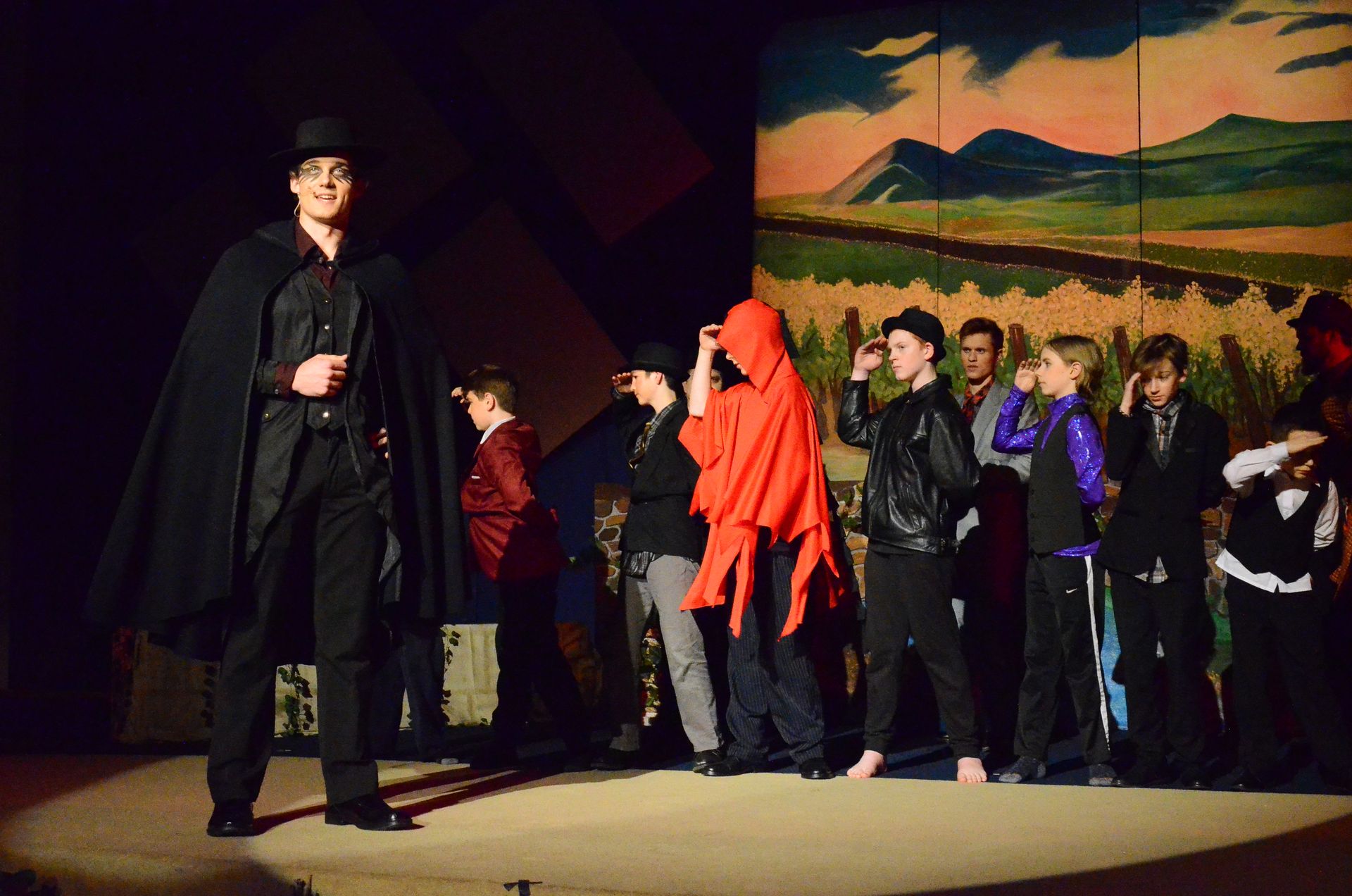 A performer in a black cape and top hat stands on stage left while a group of performers in various costumes, including one wearing a bright red cloak, gather together on a painted backdrop depicting a wheat field and distant mountains.