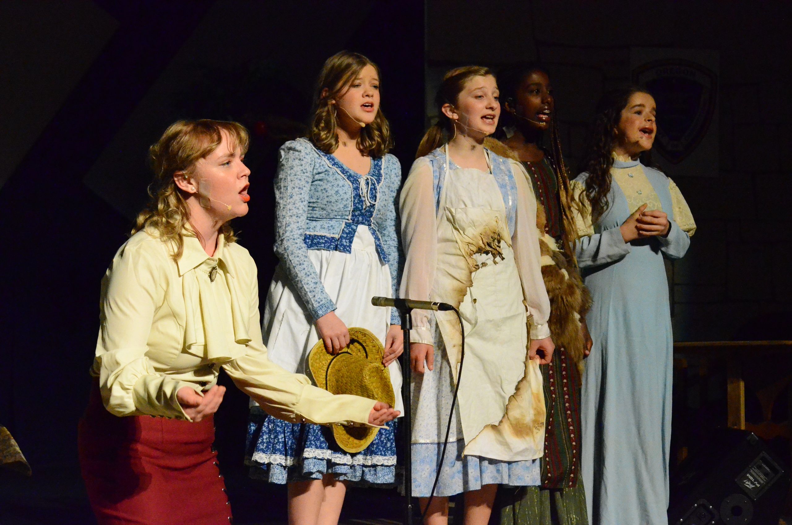 A cast of five performers in period costumes sing together on stage, with one holding a straw hat and another playing a stringed instrument, against a dark background.