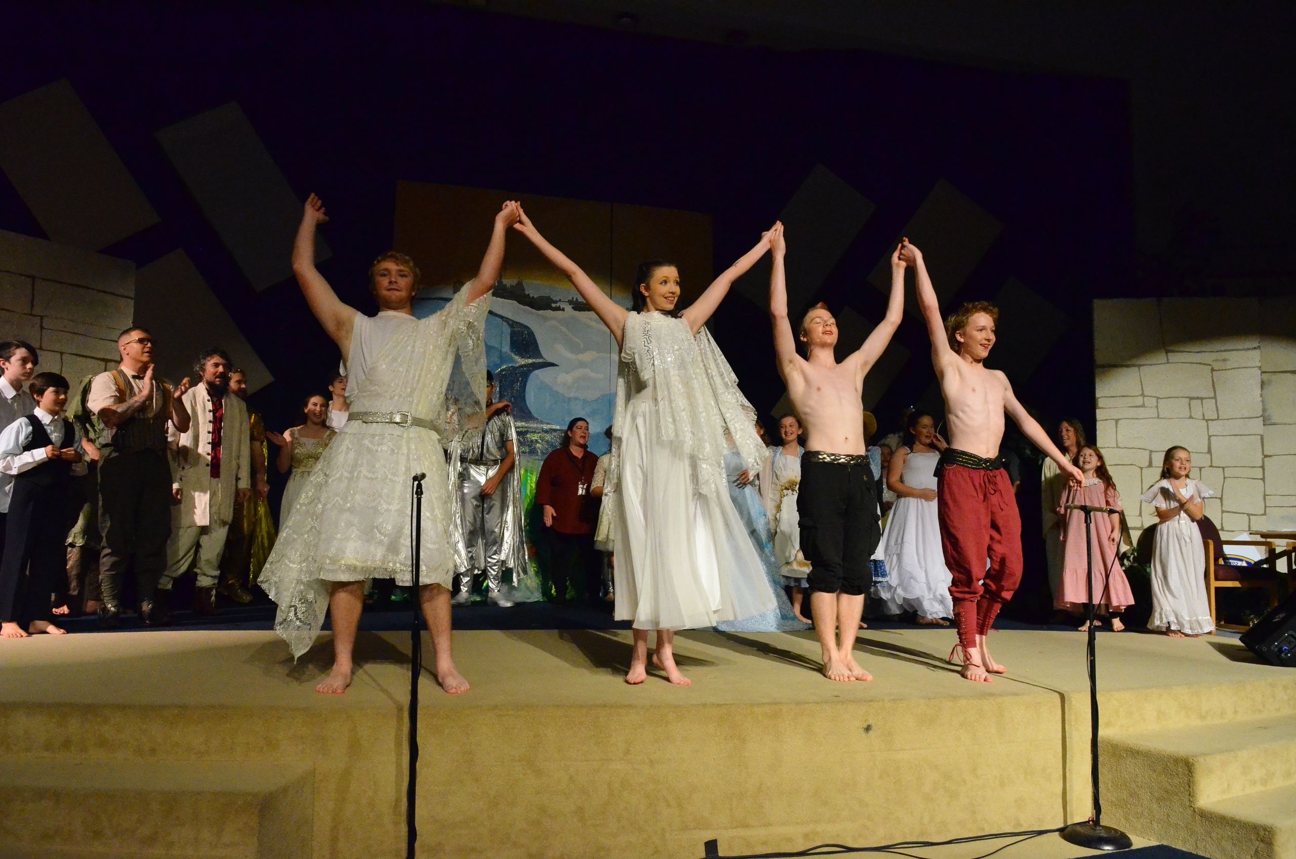 A cast of performers in classical and biblical-inspired costumes raises their hands in celebration on a raised stage set with stone walls and geometric backdrop elements, with additional cast members visible in the background.