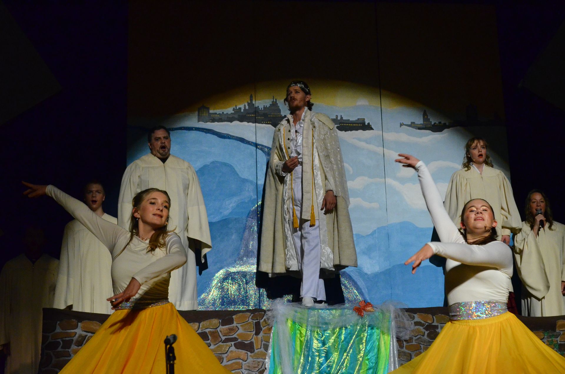 A cast of performers in cream-colored robes and yellow skirts stand on a stone-decorated stage before a backdrop depicting mountains and a distant cityscape, with the central performer elevated on a platform and others arranged around him in expressive poses.