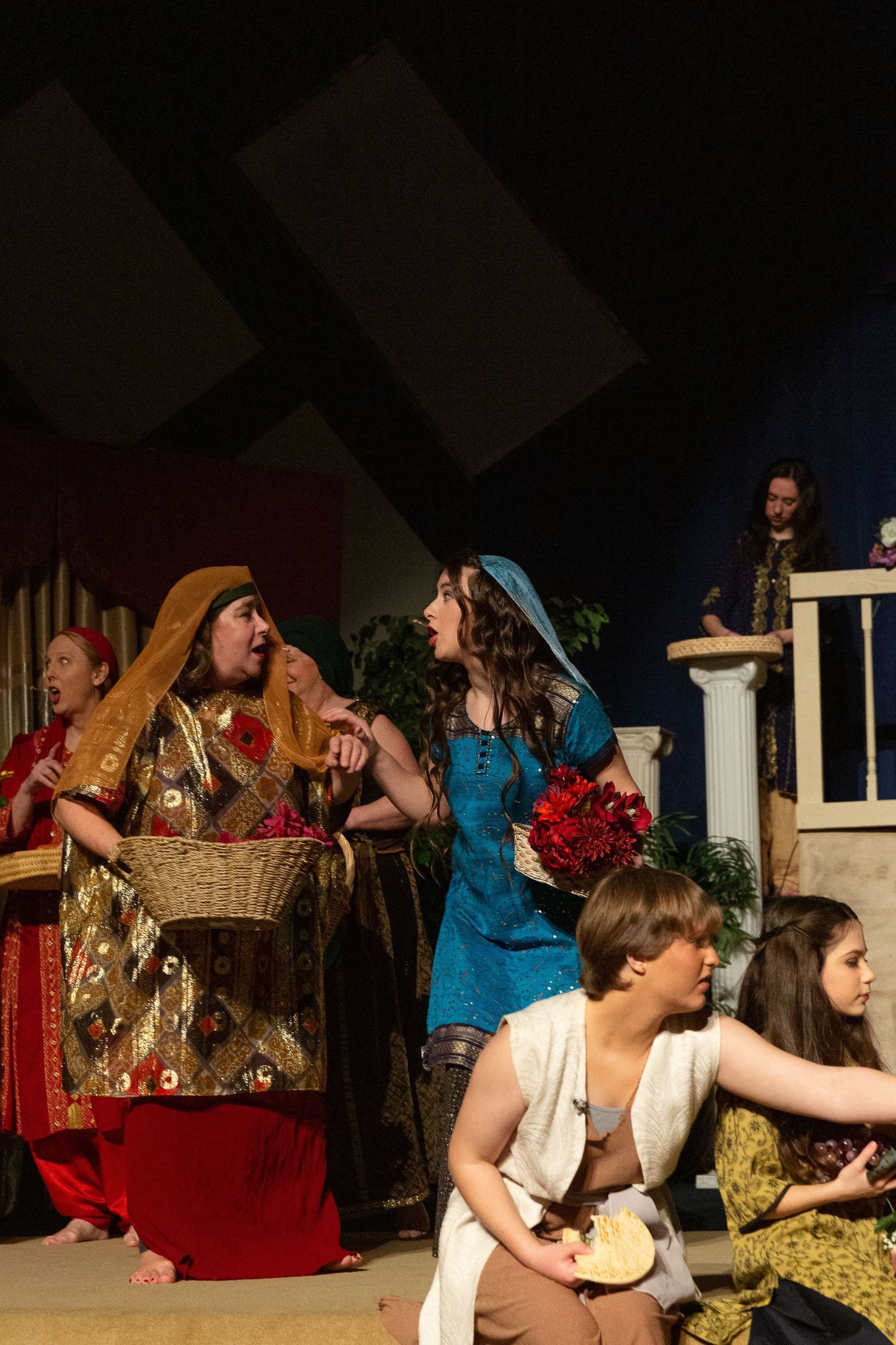 Performers in ornate period costumes engage in an animated scene on a theatrical stage decorated with classical columns and potted plants, with additional cast members visible in the background.