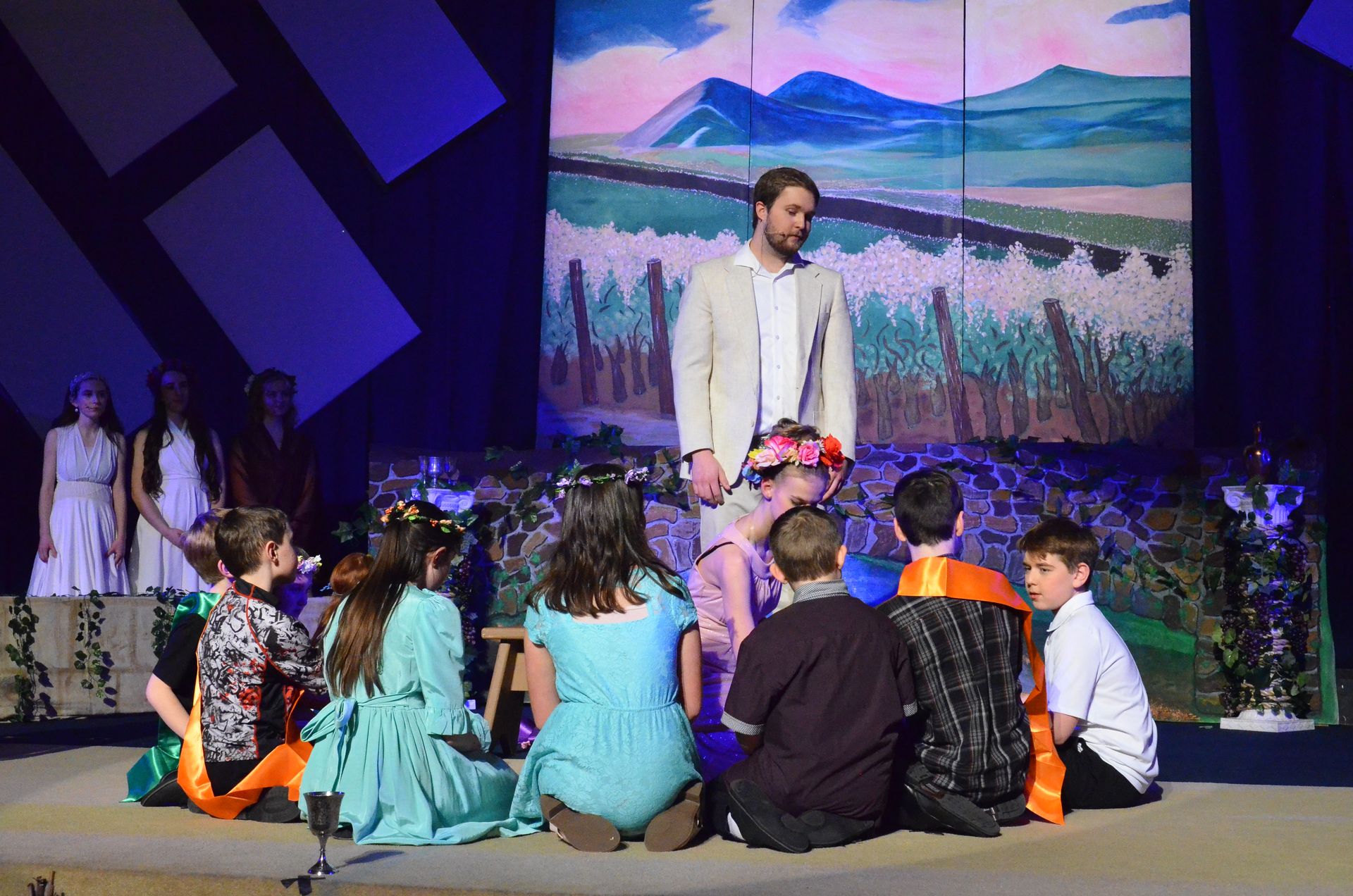 A cast of performers in colorful costumes sits on stage facing a male performer in a cream-colored jacket, with a painted backdrop of mountains and fields behind them and blue geometric set pieces flanking the scene.