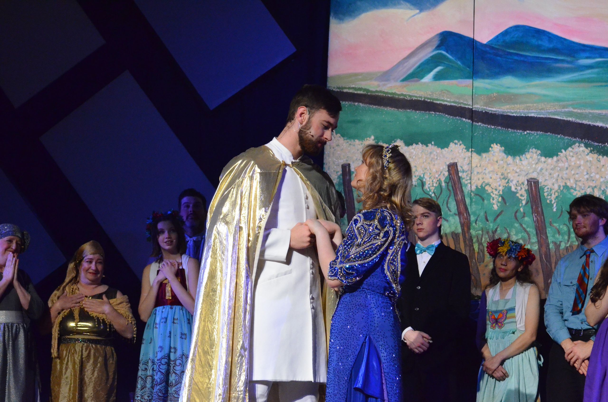 A bearded performer in a gold-trimmed white robe stands center stage surrounded by costumed cast members, with a scenic backdrop depicting mountains and a pastoral landscape.