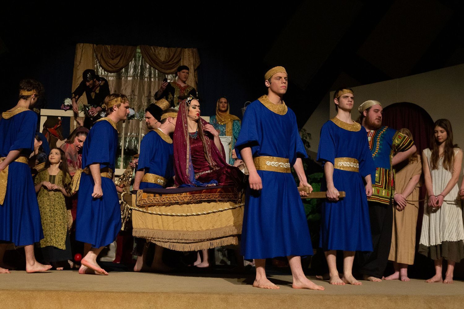 The cast performs on stage in elaborate blue tunics with gold trim and richly detailed period costumes, arranged in a formal tableau scene with ornate draped fabric backdrops and theatrical set pieces.