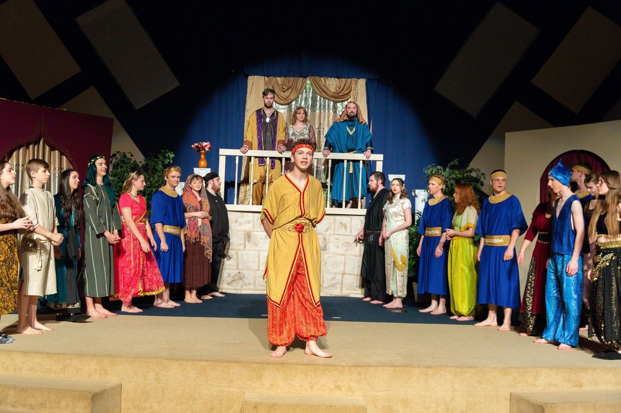 A cast of performers in colorful robes and tunics stands on a multi-level stage set with classical columns and draped gold curtains, with the central performer in a yellow and red garment striking a dramatic pose while ensemble members surround the scene.